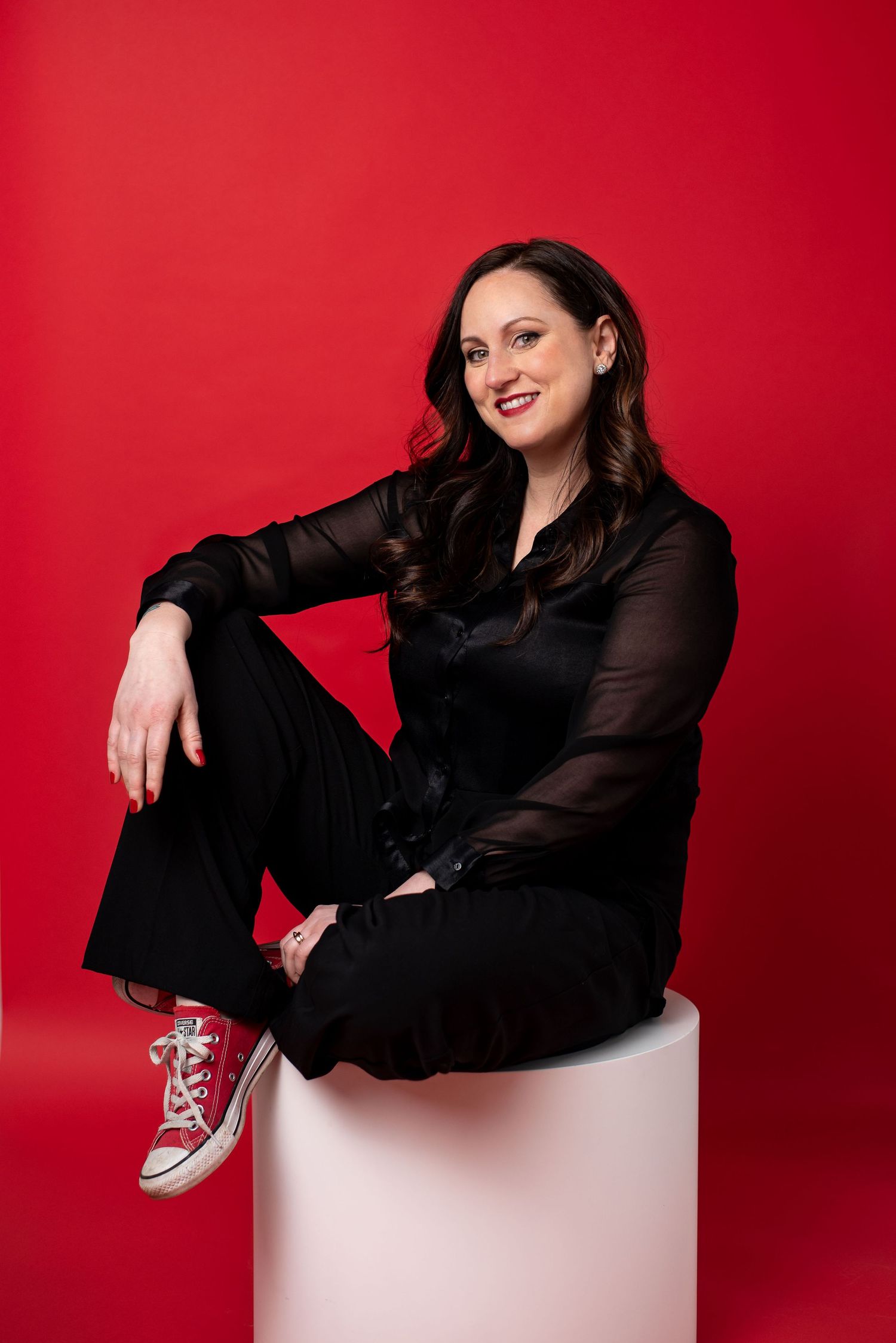 Portrait of person in black outfit sitting on white pedestal against vibrant red background wearing casual sneakers.