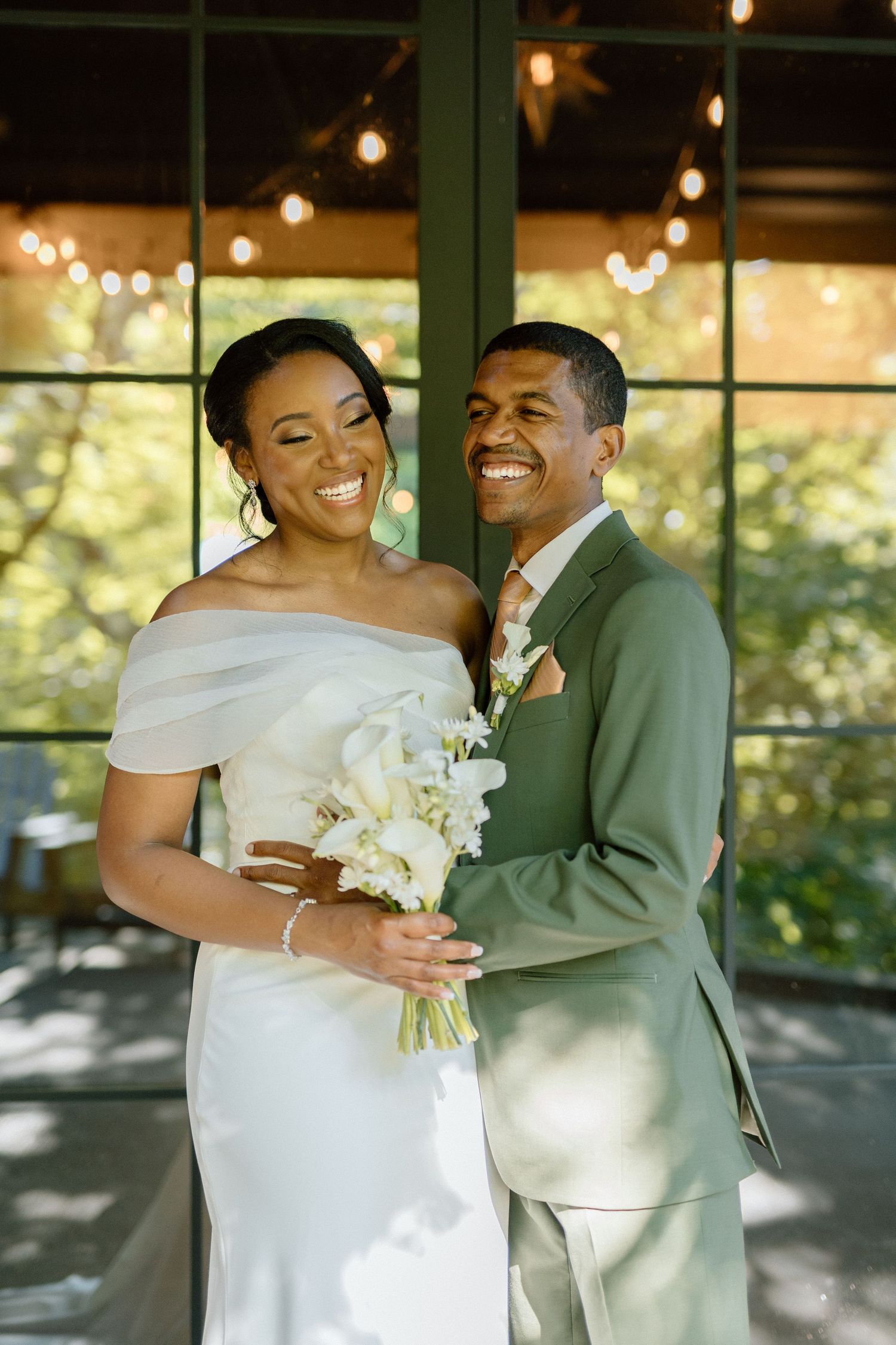 A joyful couple shares a laugh on their wedding day, with the bride in white and groom in sage green against string lights.