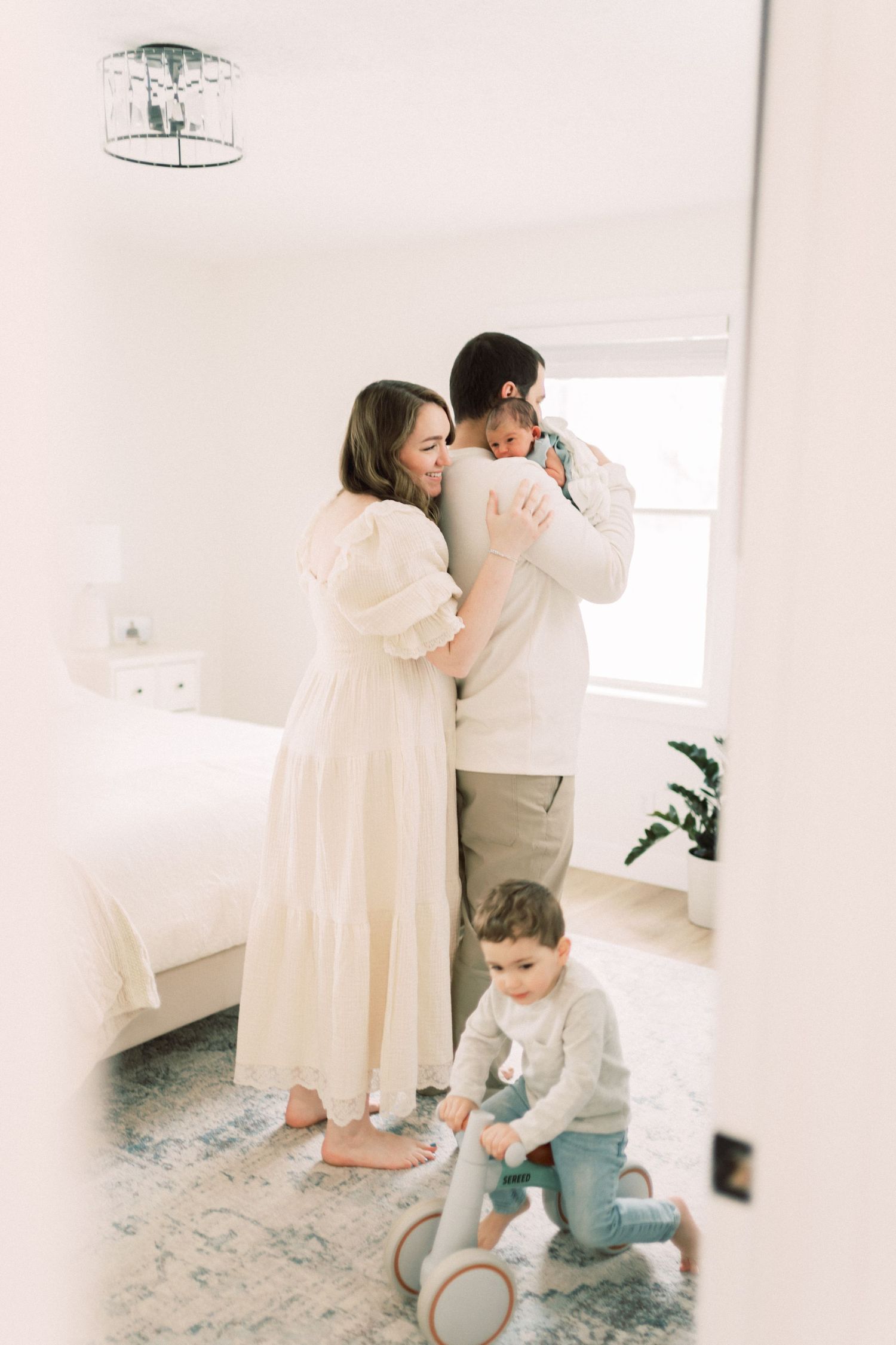 Family in soft white attire sharing a tender moment in bright minimalist room while toddler plays with toy scooter.