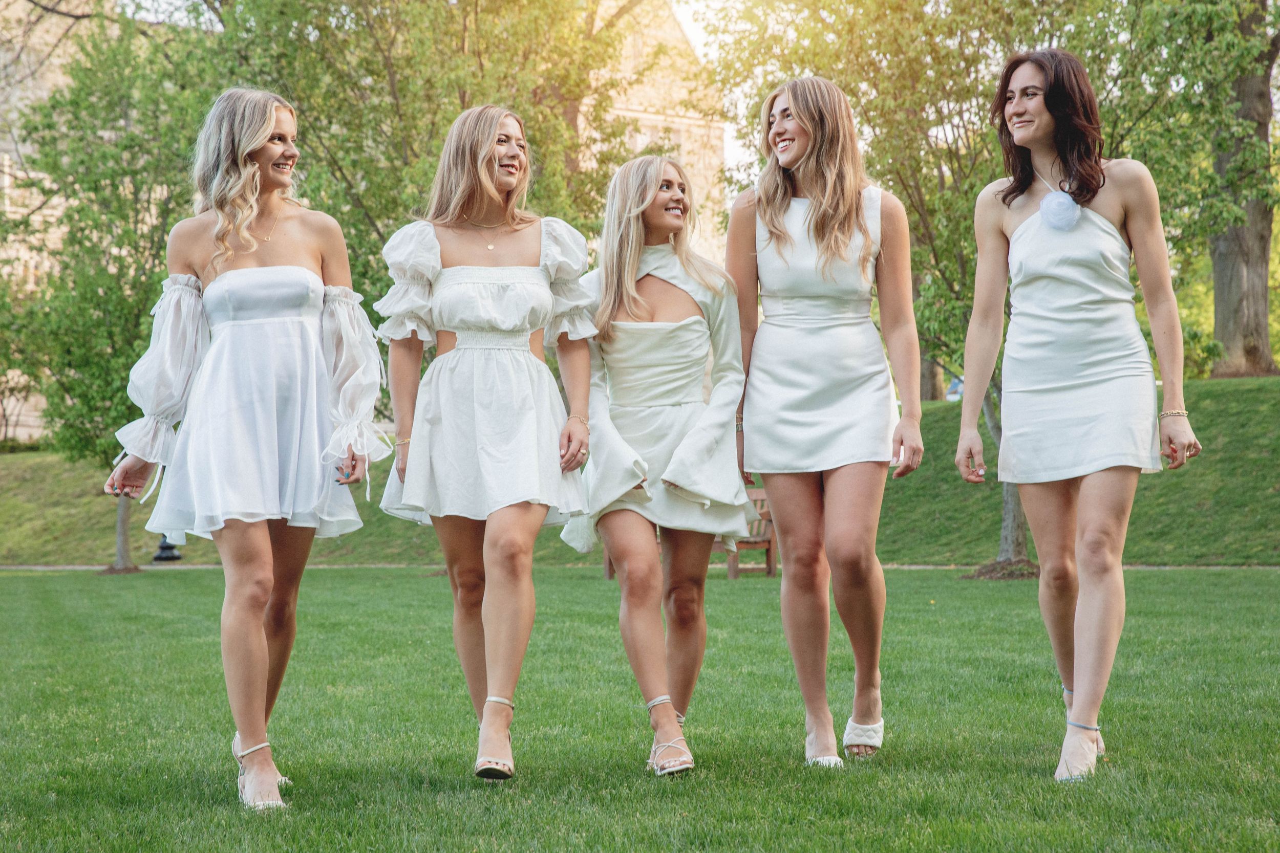 Five people wear matching white summer dresses as they walk together across a sunlit lawn.