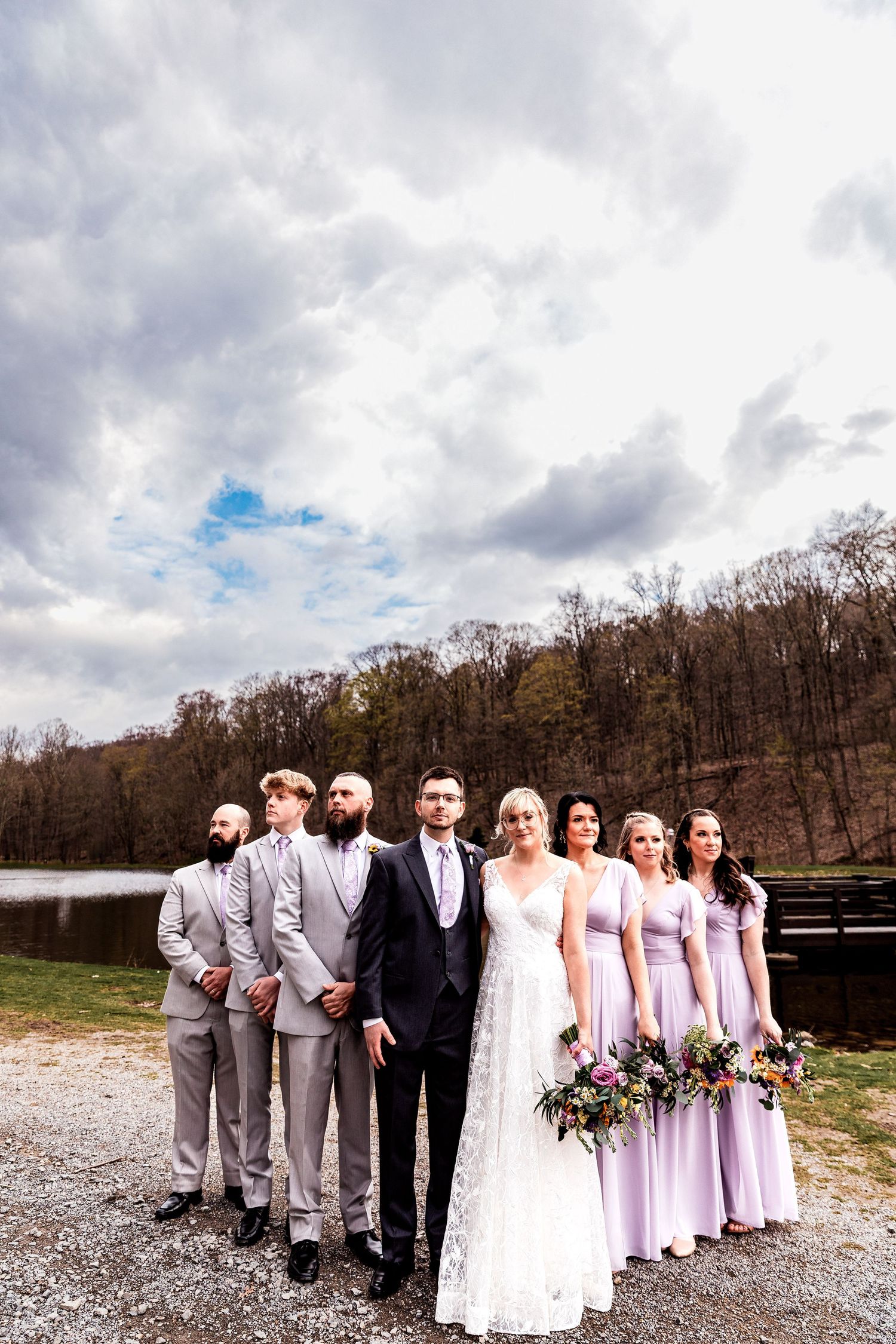 A wedding party in grey and lavender attire poses together by a lake on a cloudy day with bare trees in the background.