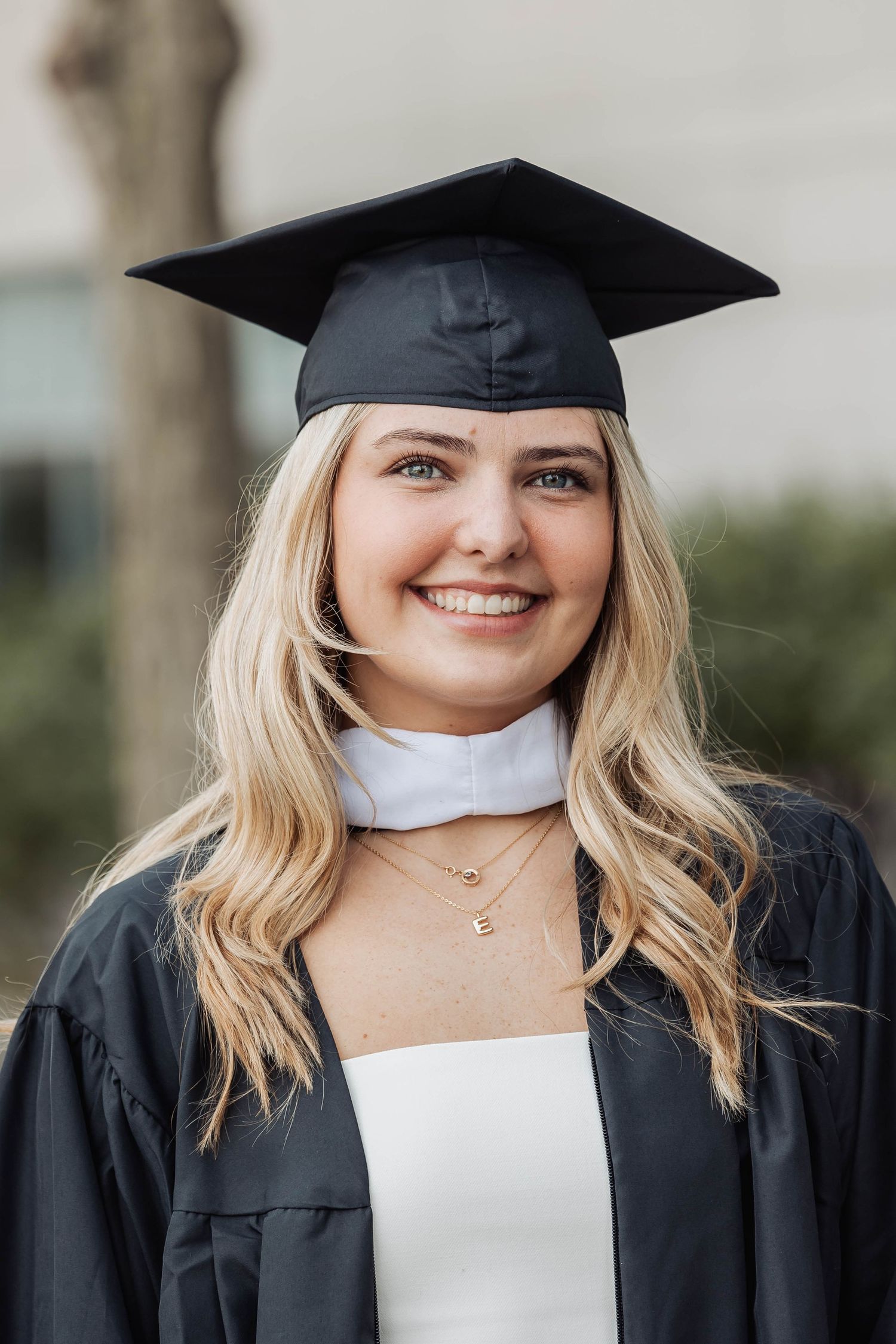 A graduate in traditional black cap and gown with white collar smiles outdoors in a graduation portrait.