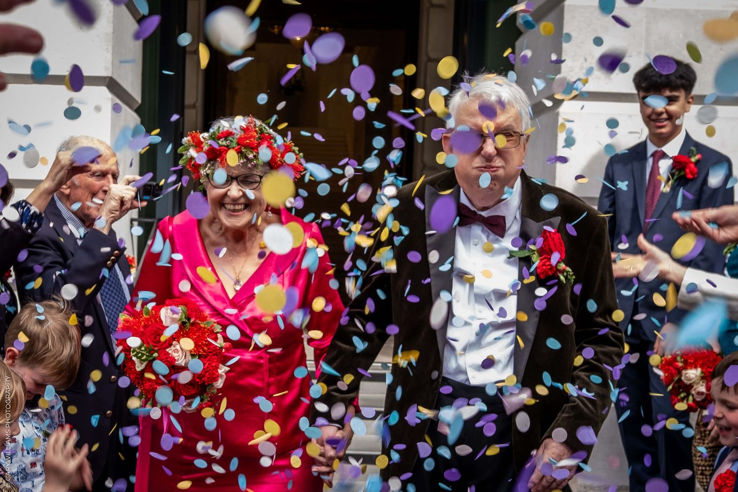 A festive celebration with colorful confetti falling while people in bright attire stand outside a building entrance.