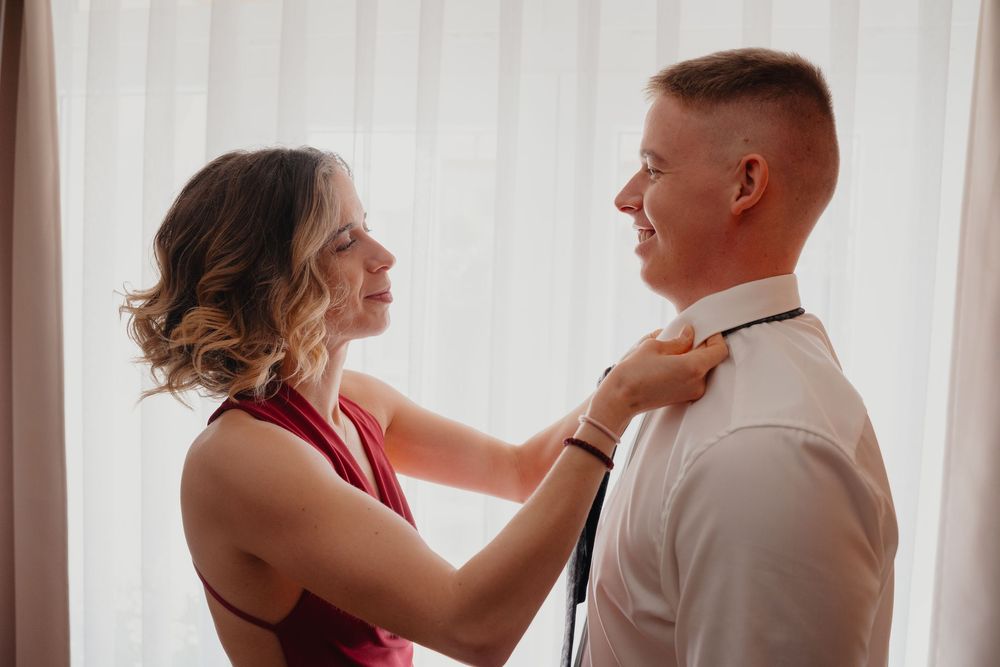A person in a burgundy dress helps adjust a tie on someone wearing a white dress shirt against a curtained background.