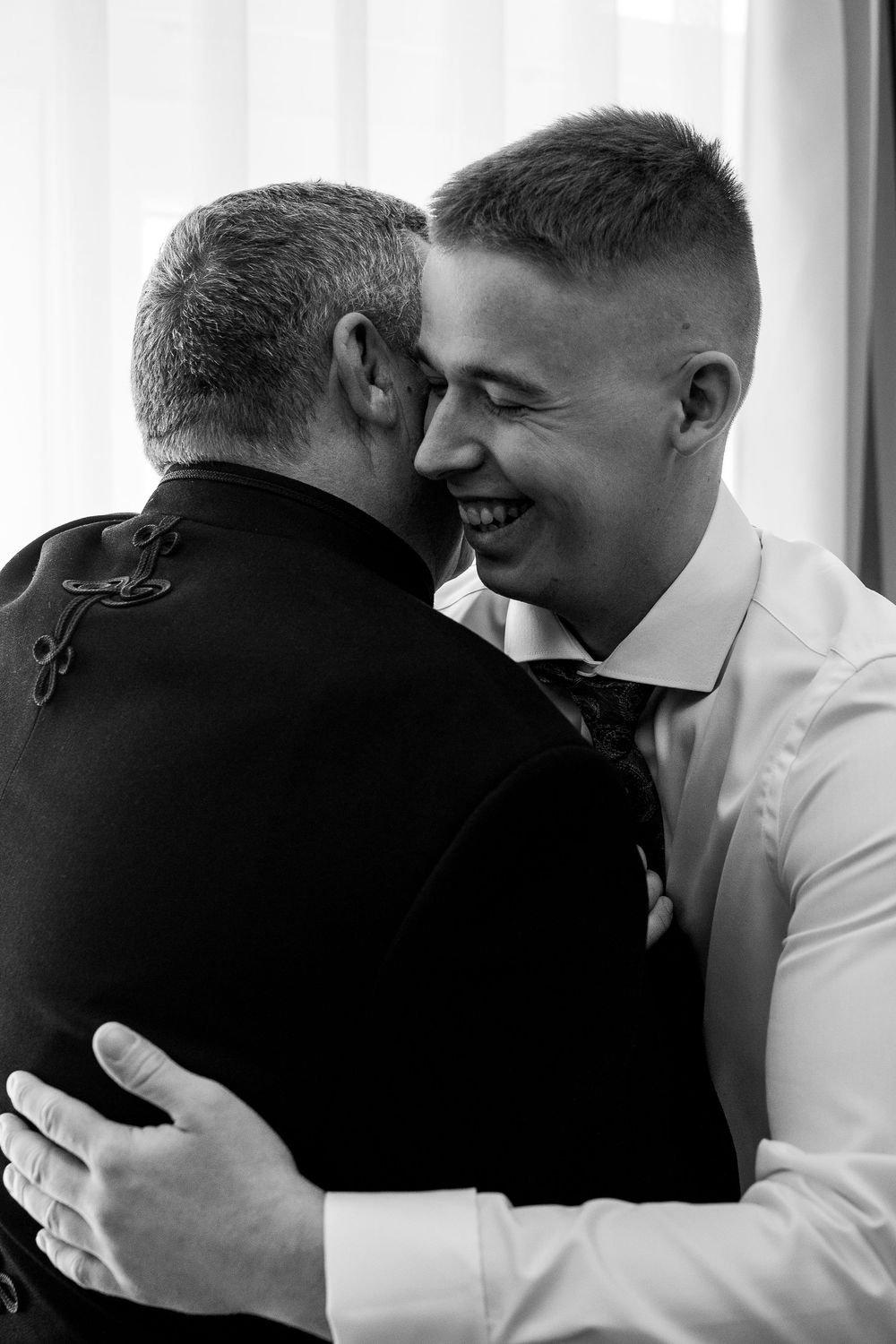 Black and white photo capturing an emotional embrace between two men in formal attire, one smiling warmly.