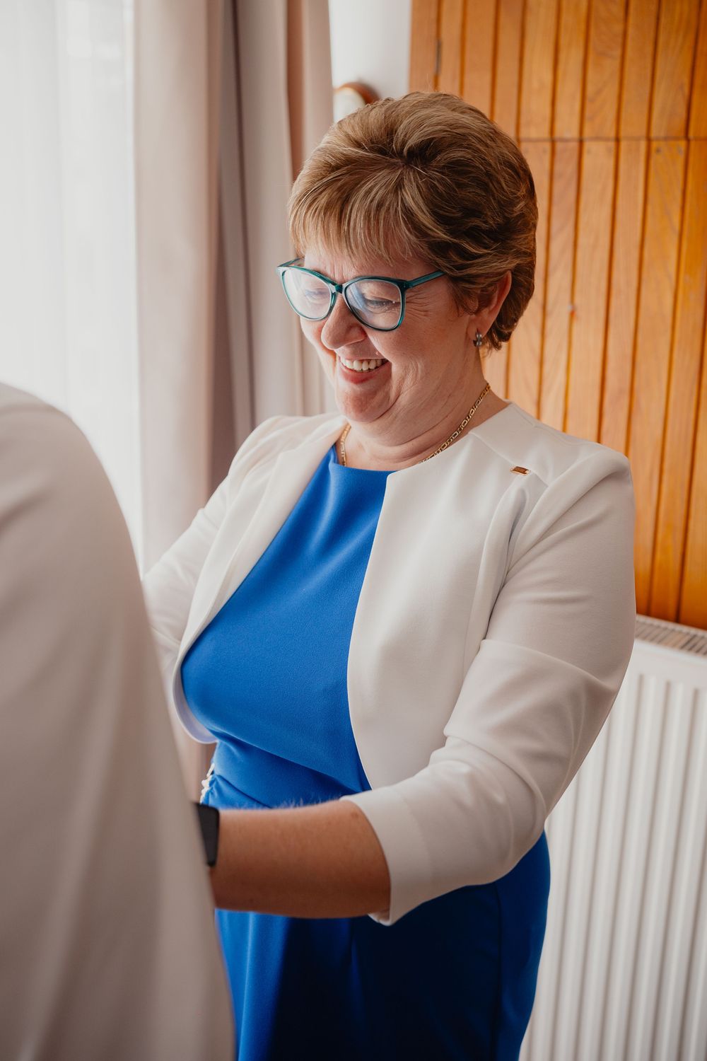 A person wearing glasses and a blue dress with a white cardigan laughs joyfully while standing near a window.