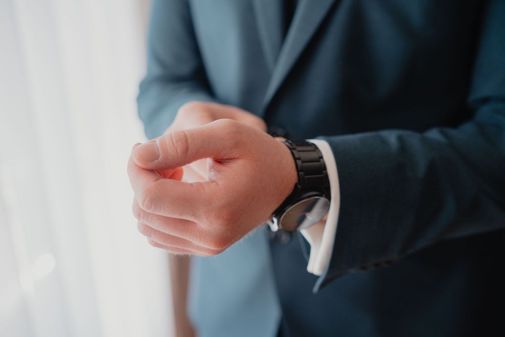 Close-up view of hands wearing a formal suit sleeve checking a metallic wristwatch against white background.