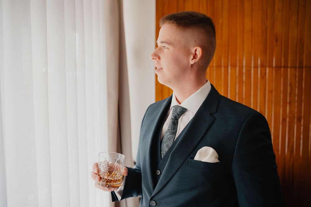 A person in a dark suit and tie holds a whiskey glass while standing by a window with wooden paneled walls.