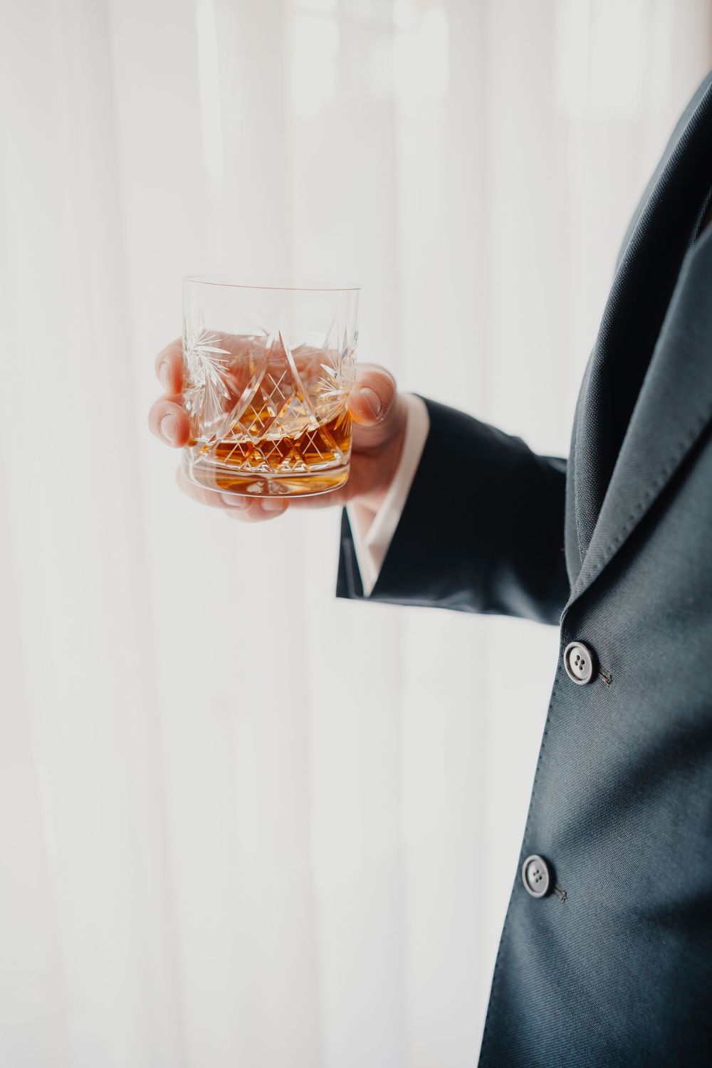 Hand in formal black suit jacket holds a crystal glass of whiskey against a white background.