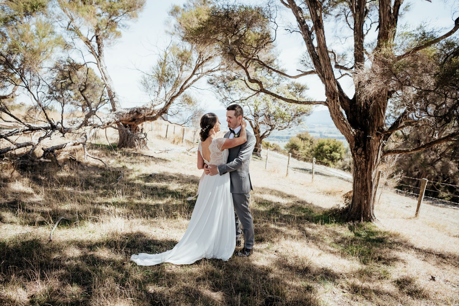 A couple embraces under tall Manuka trees on a sun-dappled hillside at Whitimanuka during an outdoor wedding ceremony by August and Vine