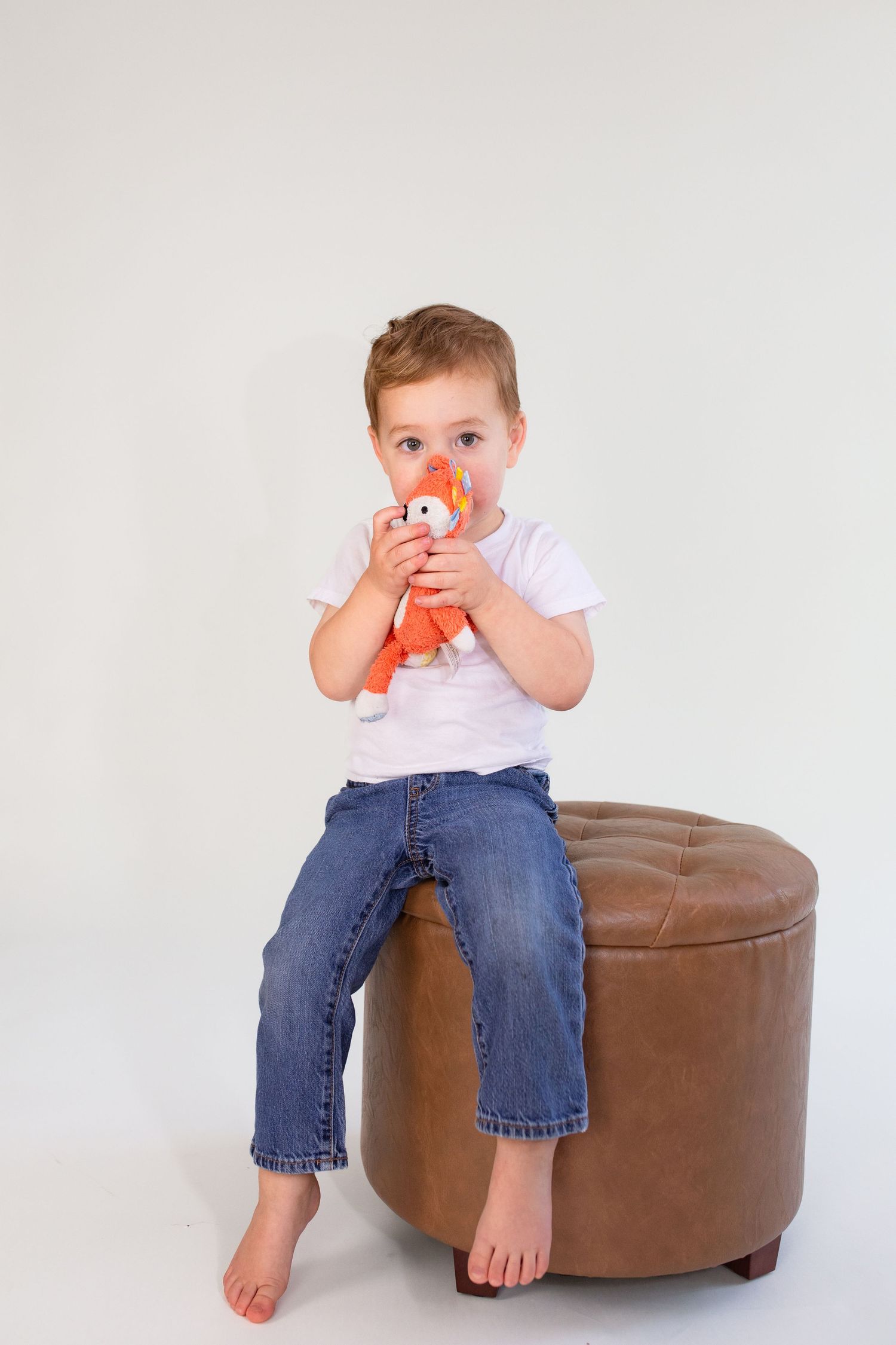 A small child in jeans sits barefoot on a brown leather ottoman while holding a plush fox toy.