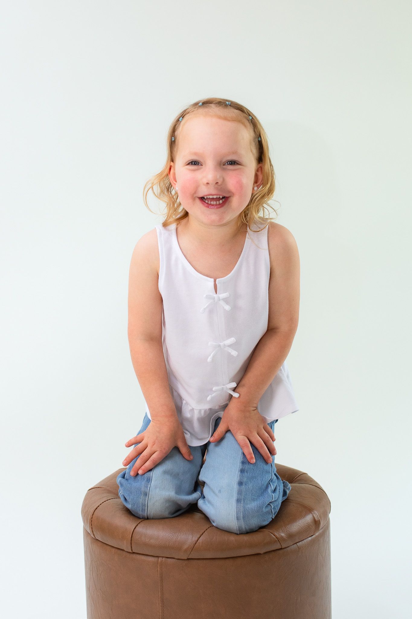 Young child in white tank top and jeans kneels on brown leather ottoman while laughing joyfully against white background.