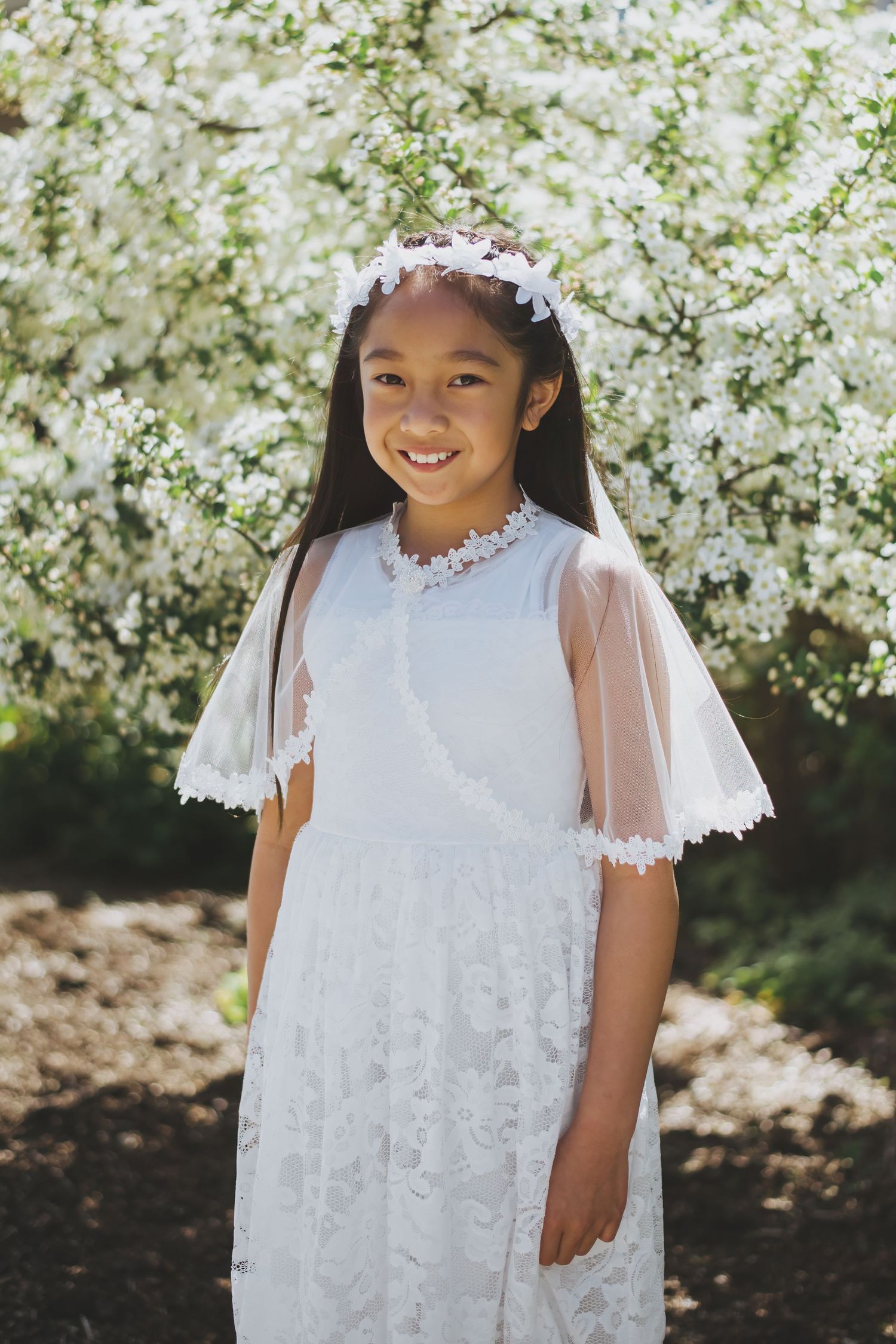 A child wearing a white lace dress and floral headpiece stands in front of blooming white flowers in a garden setting.