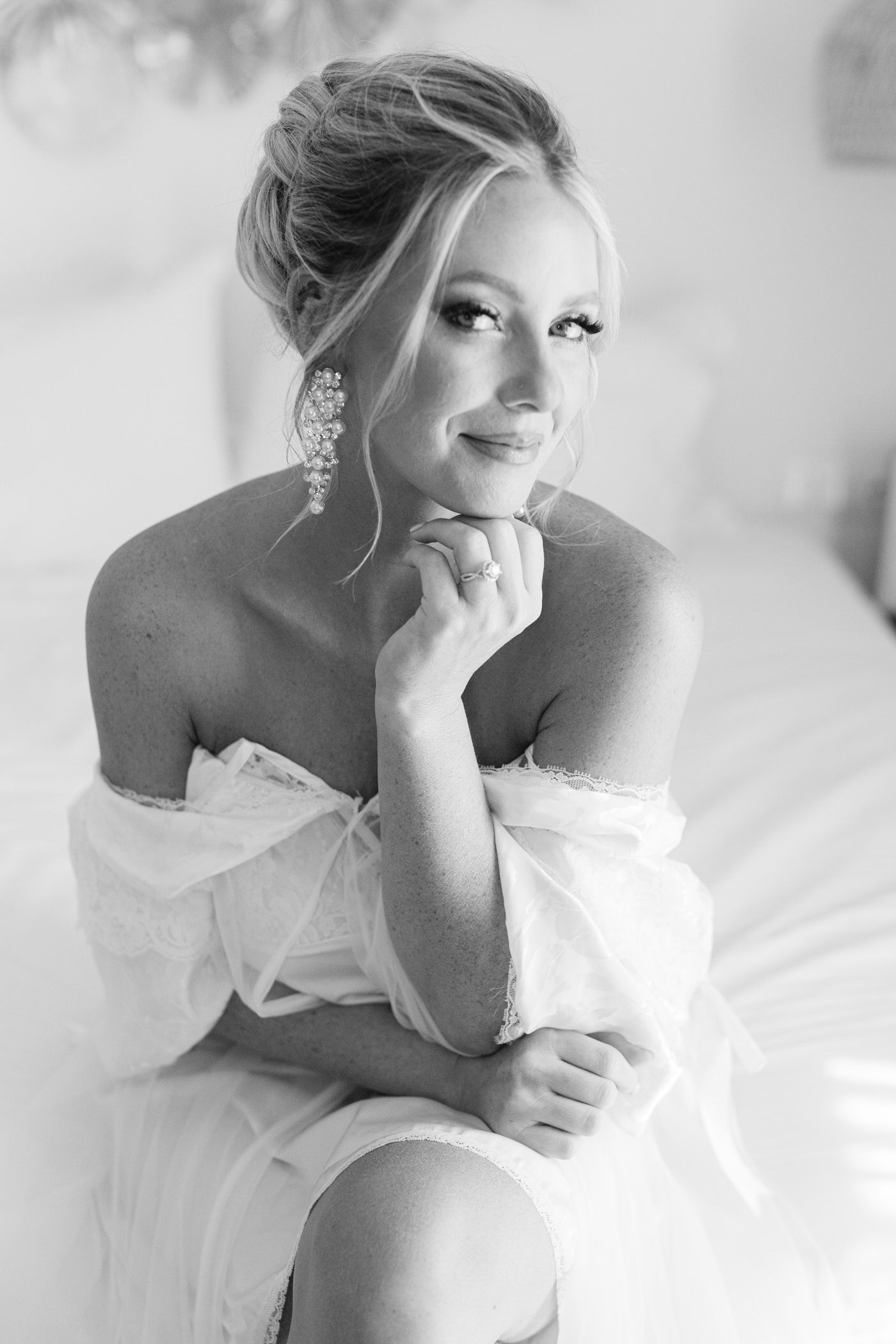 Black and white portrait of a bride in an off-shoulder gown with statement earrings posing elegantly with hand on chin.