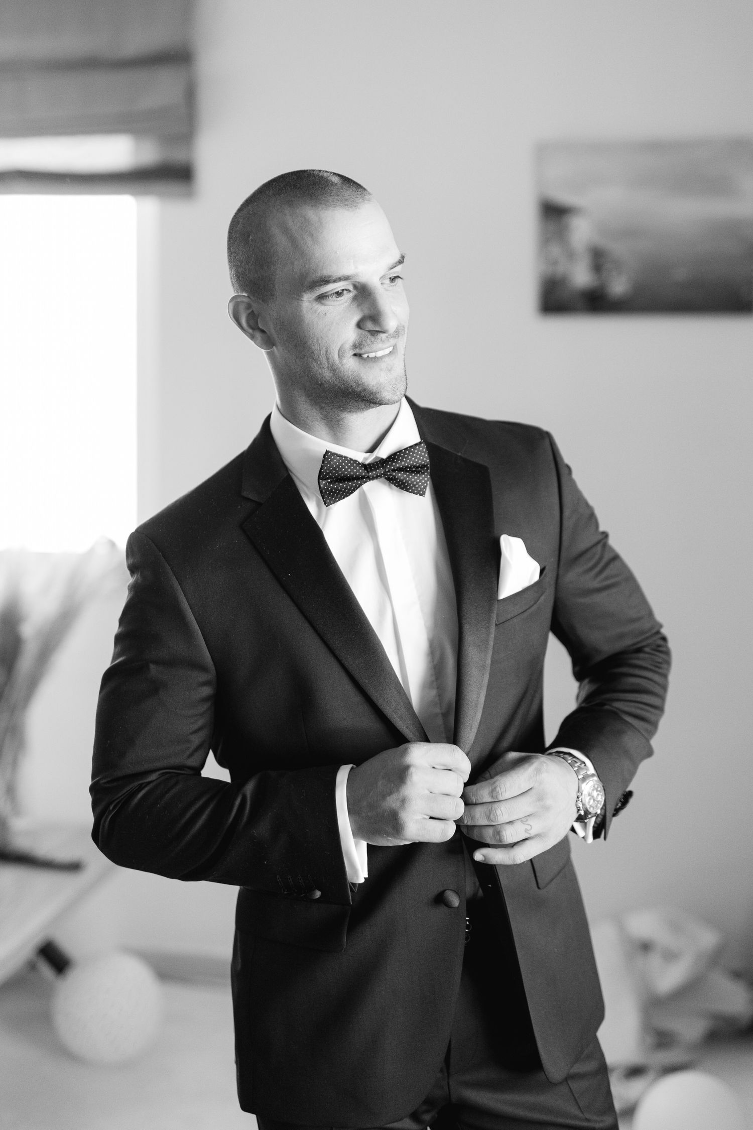 Black and white photo of person in classic tuxedo with bowtie adjusting jacket buttons while smiling.