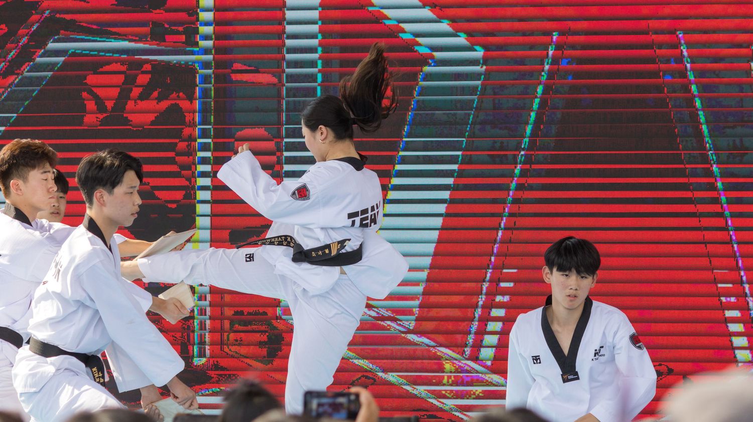 Taekwondo athletes practicing martial arts moves with kicks and strikes against a vibrant red stadium backdrop.
