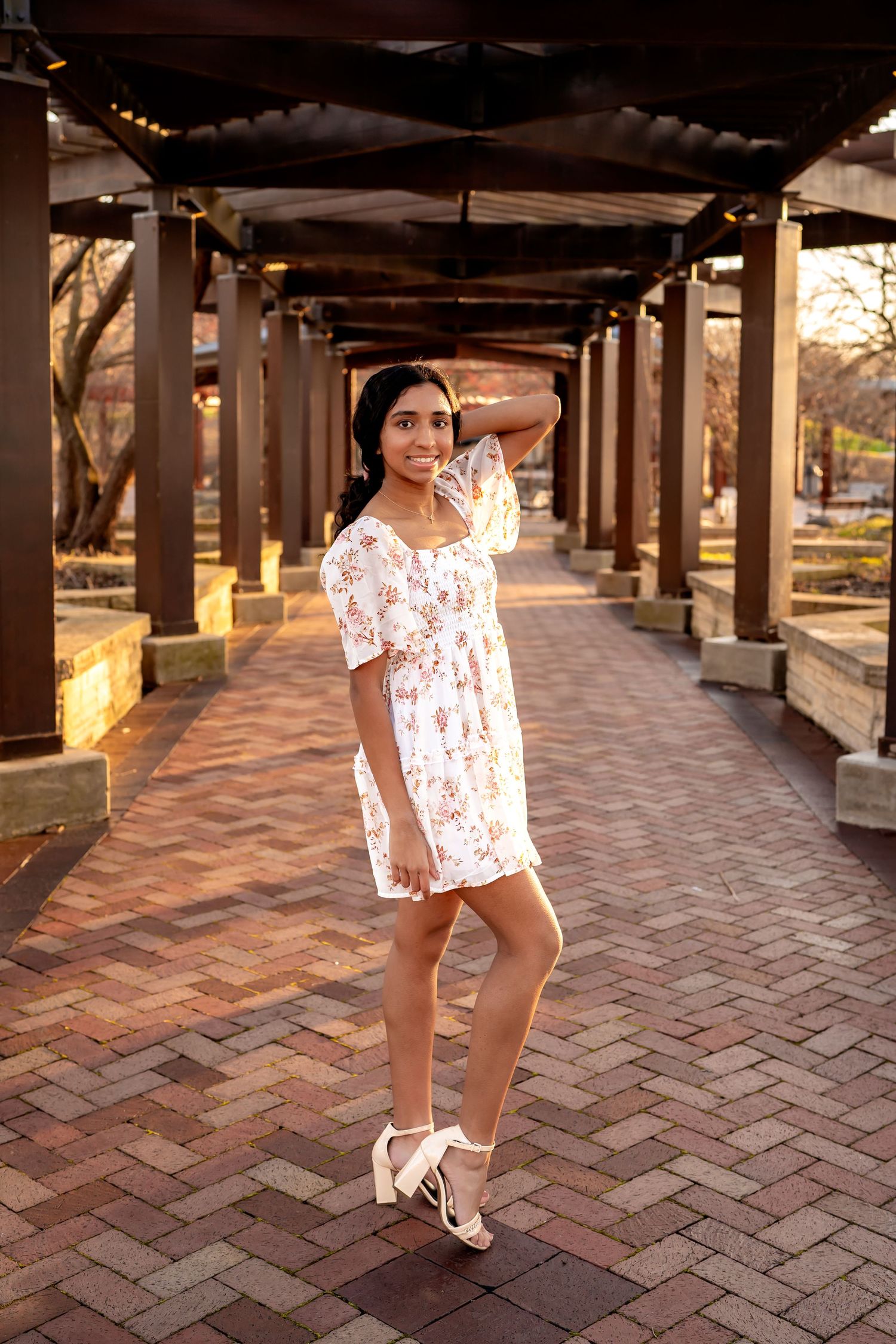An elegant figure poses in a floral white dress and heels under a covered brick walkway during golden hour.
