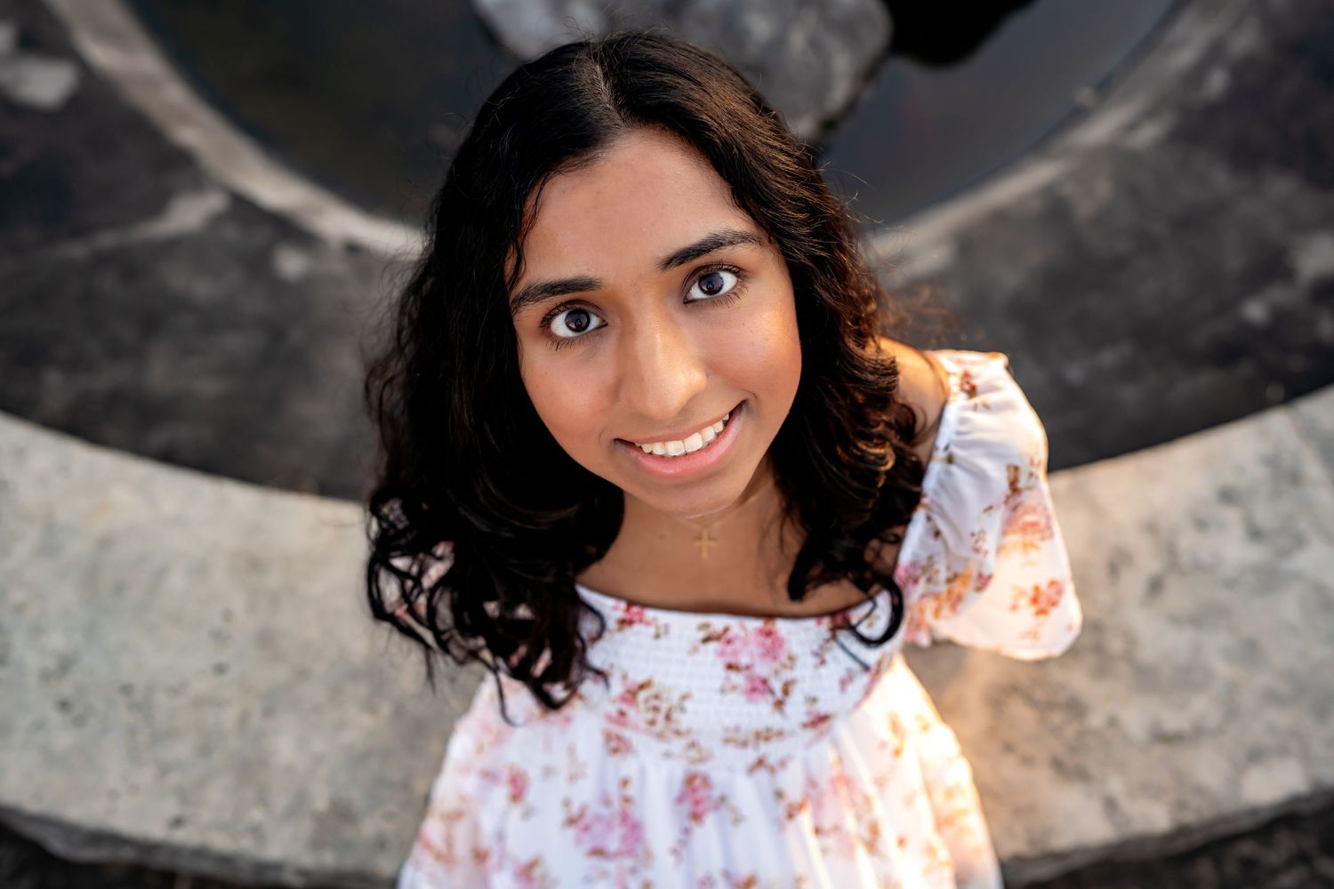 A close up portrait shot featuring a warm smile and dark curly hair, wearing a white floral dress in natural lighting.