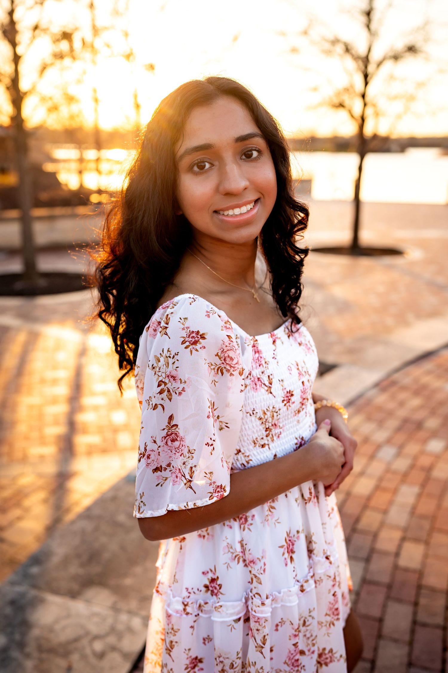 Young woman in white floral dress smiles during golden hour sunset by waterfront brick path.