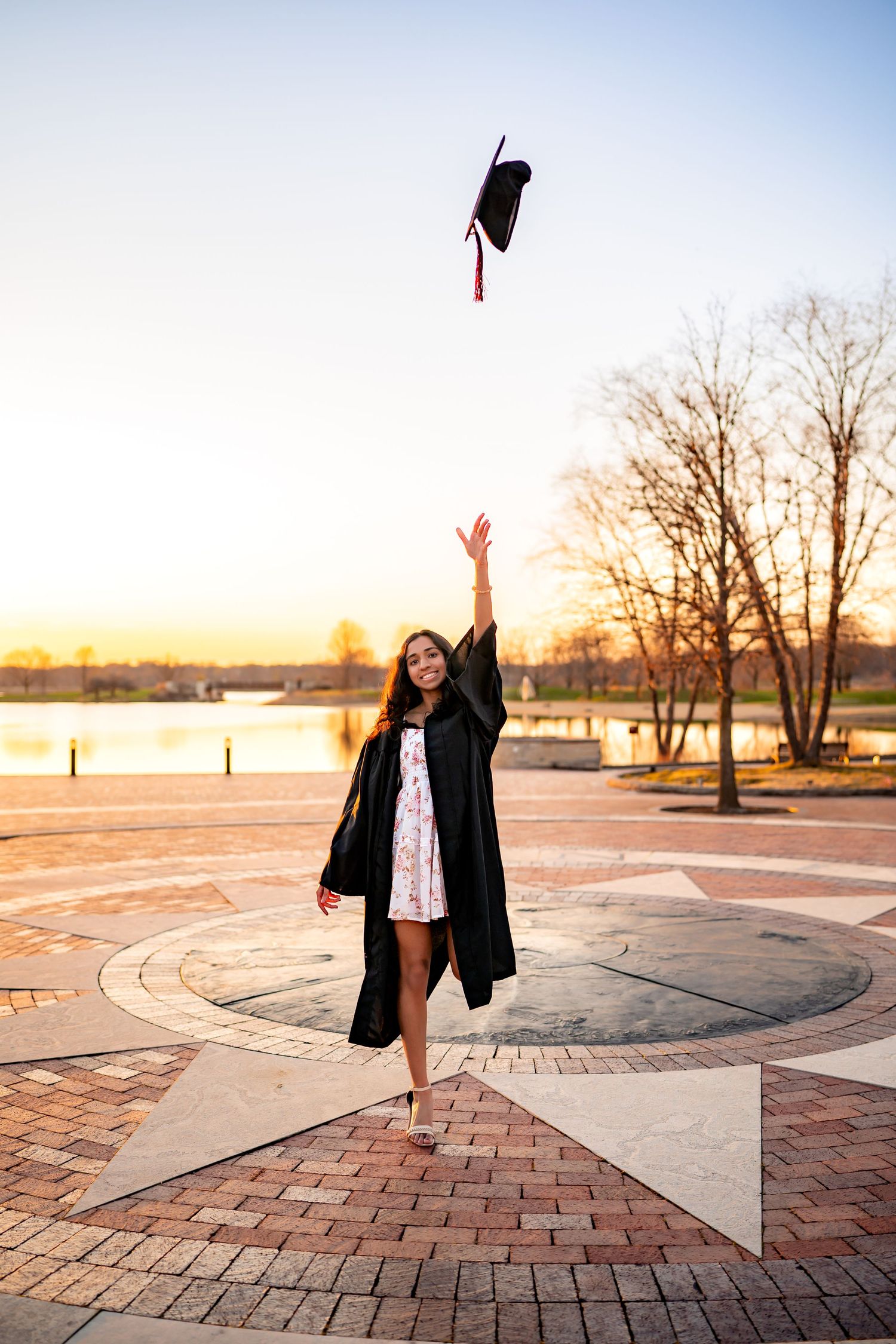 Graduate celebrating on a brick compass rose patio near water at sunset while tossing cap in the air.