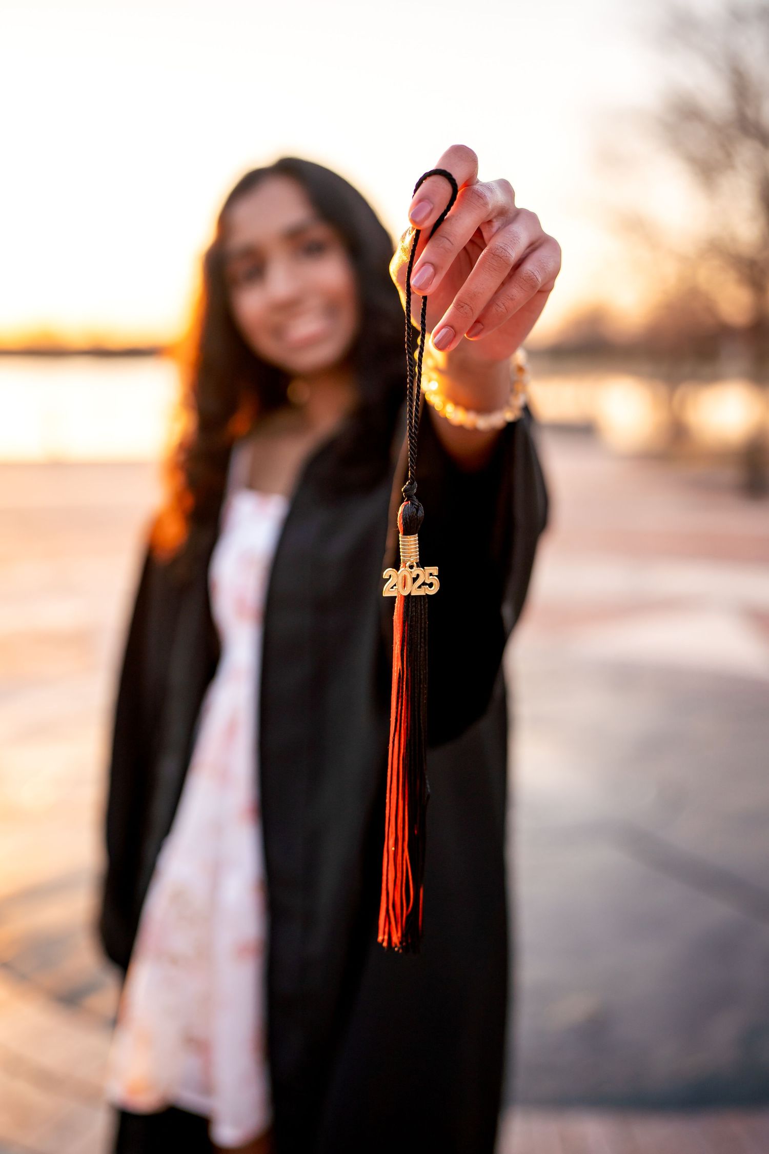 Graduate holds up red and gold graduation tassel against warm sunset backdrop in outdoor setting.