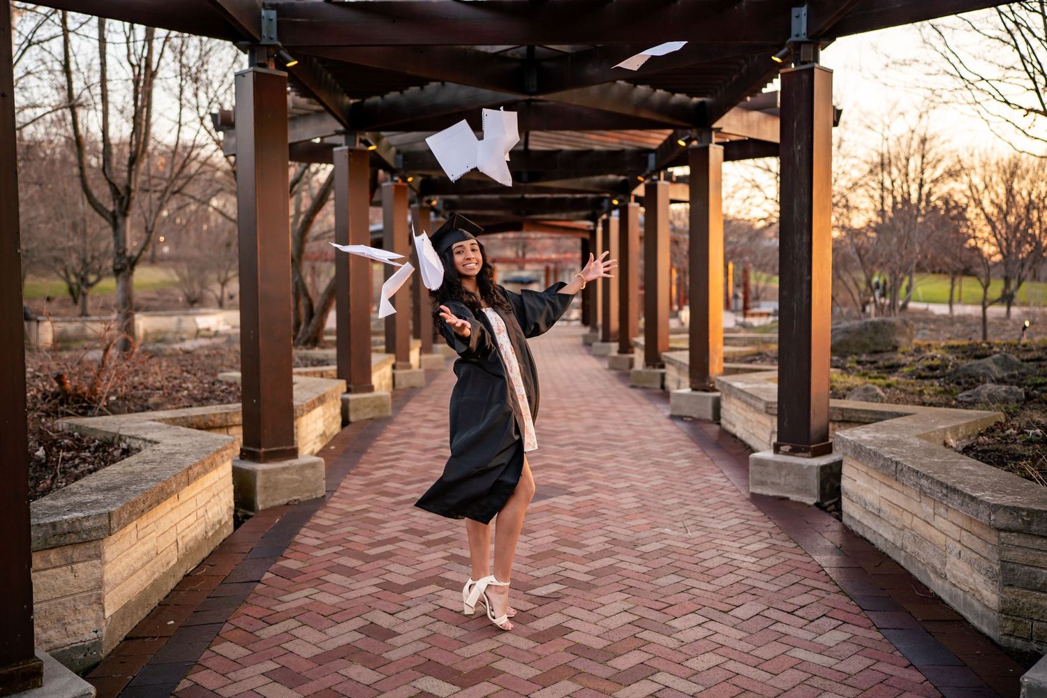 A graduate celebrating by tossing papers in the air under a covered brick walkway at sunset.
