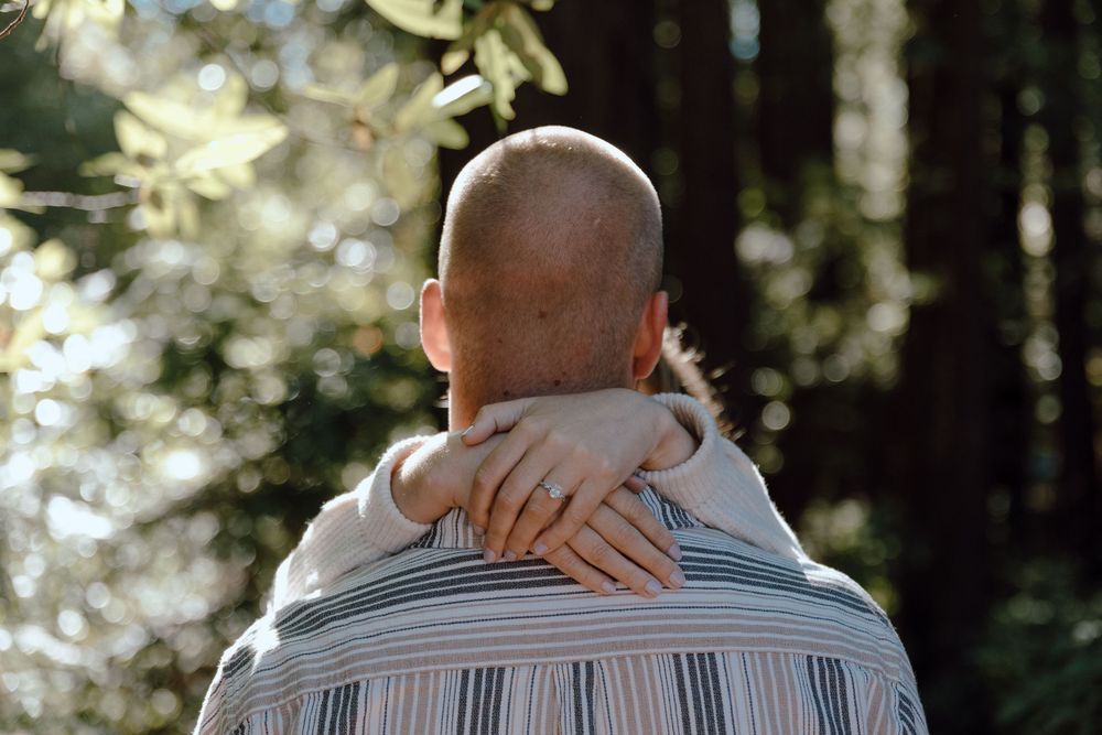 Two people embracing in a tender hug against a sunlit forest backdrop, with one wearing a striped shirt.