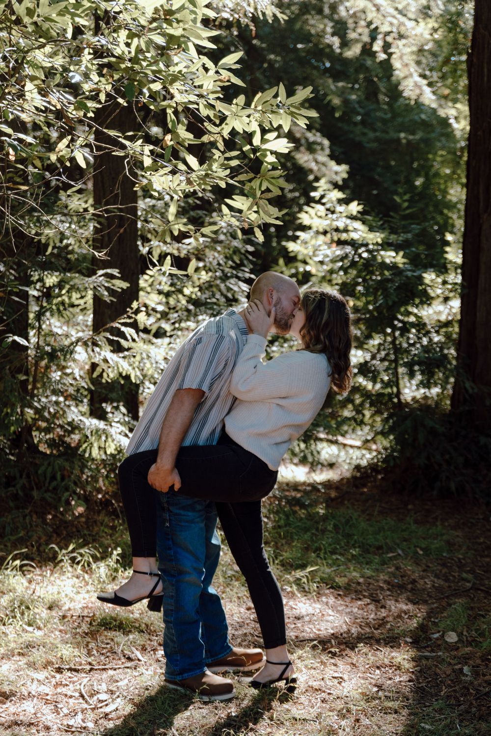 A romantic couple shares a tender kiss in a sunlit forest setting surrounded by lush greenery and dappled light.