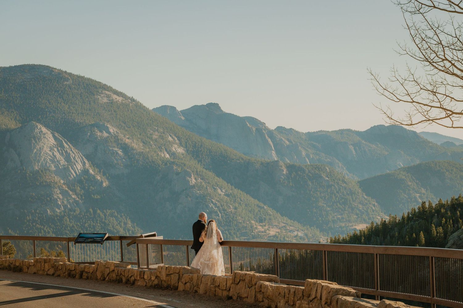 Couple admires stunning mountain vista from wooden deck observation point at scenic overlook during golden hour.