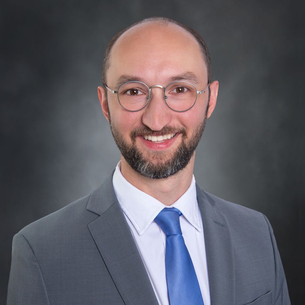 Professional headshot of a person wearing a navy blazer and white top against a gray background.