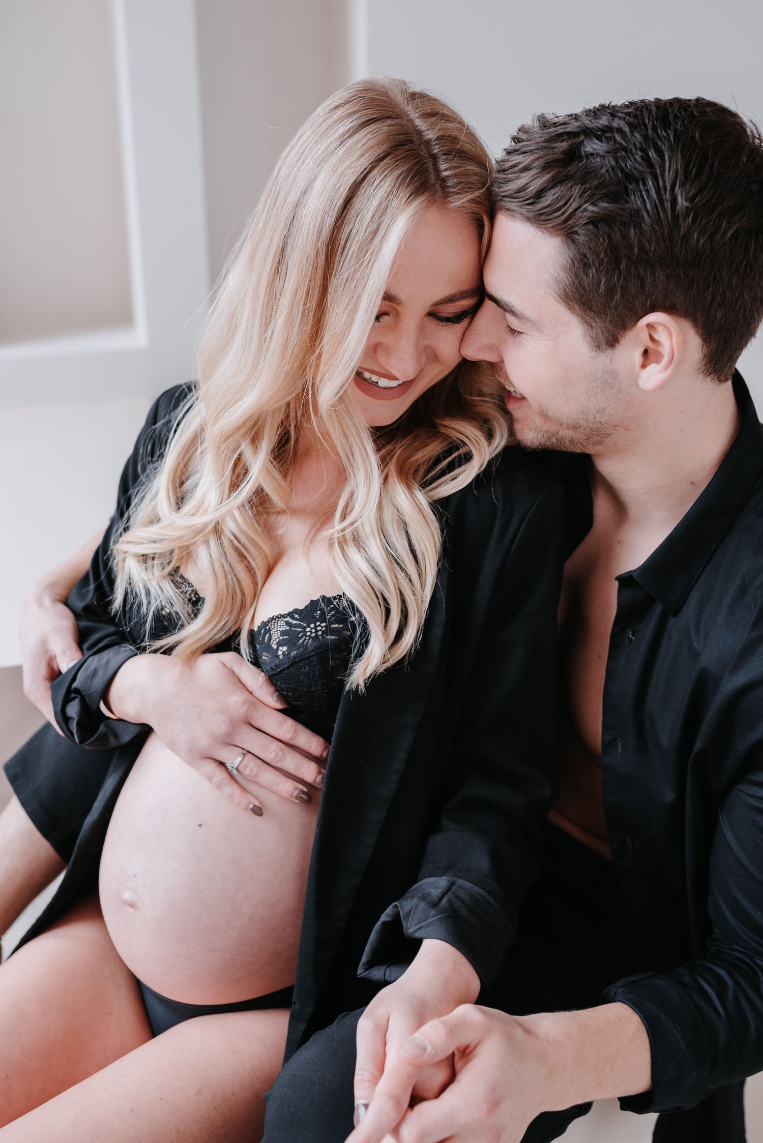 Intimate maternity photo of couple in black clothing sharing tender moment against white wall.