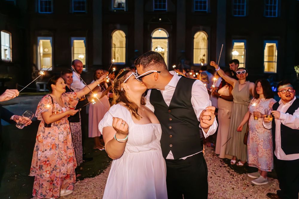 Wedding guests dance together in an elegant ballroom with arched windows and rose petals scattered on the floor.