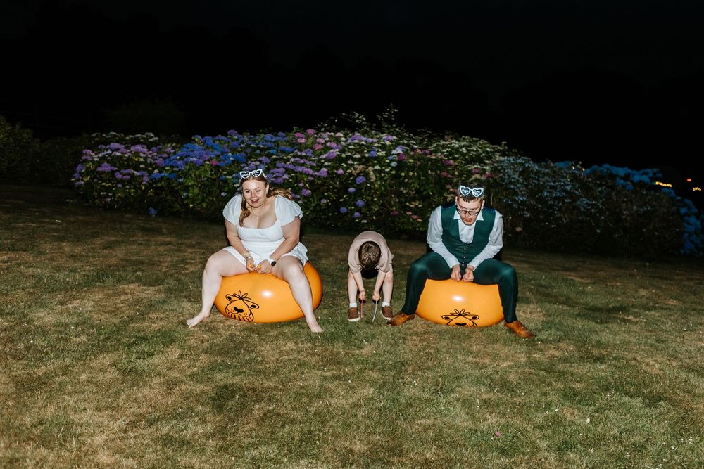 Two people sitting on orange pumpkins on a lawn with a dog between them and flowering bushes in the background.