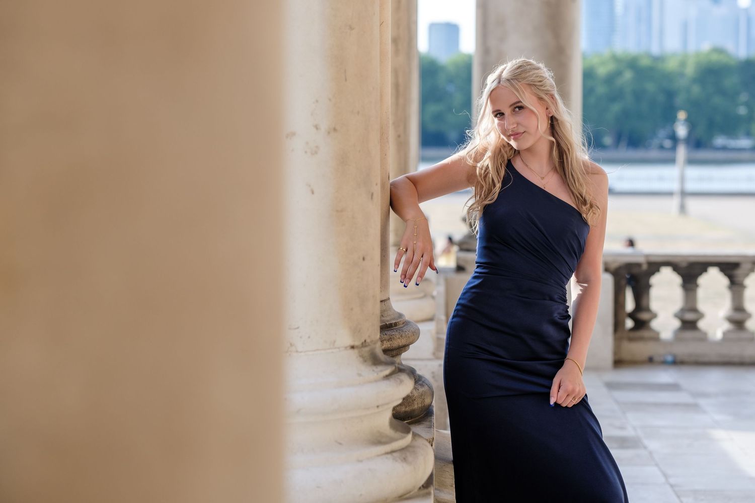 Woman in elegant black dress poses against stone column on balcony with city skyline in background.