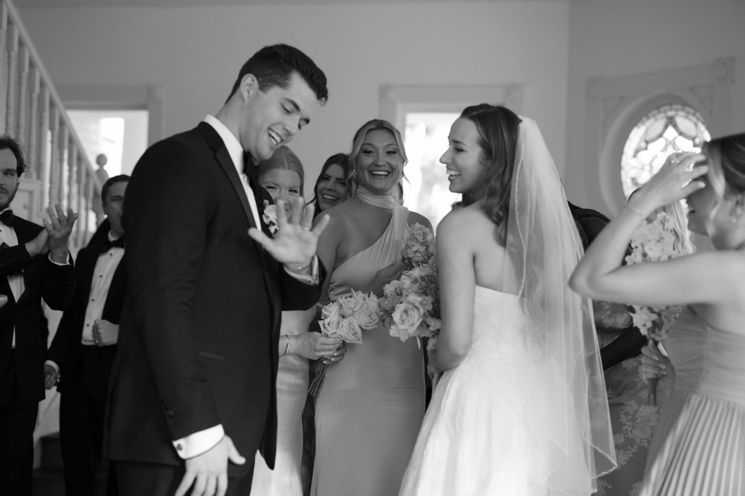 A candid black and white wedding moment shows joyful interactions between wedding party members in a bright hallway.