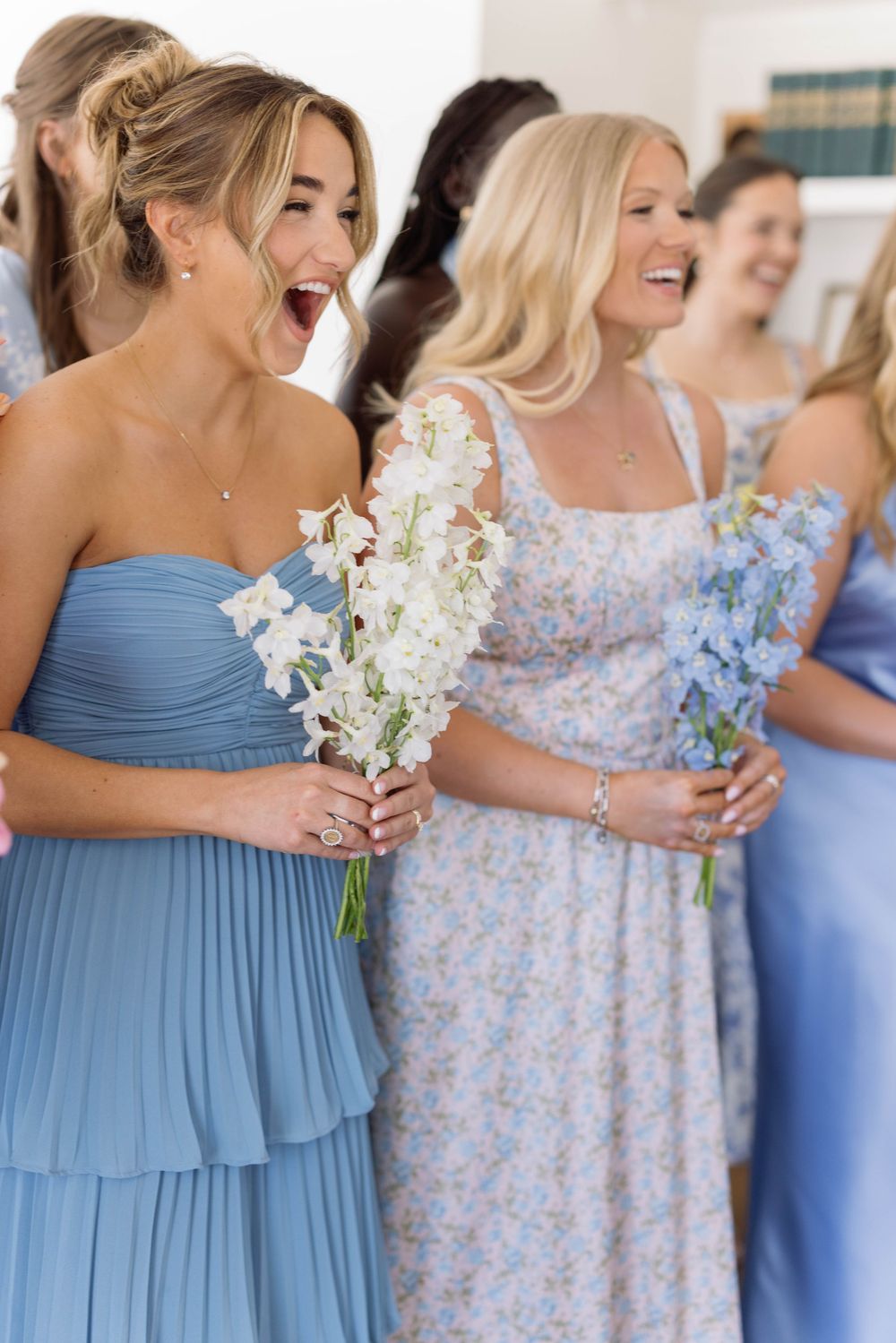Bridesmaids in blue dresses and floral prints laugh together while holding white bouquets during a wedding party.