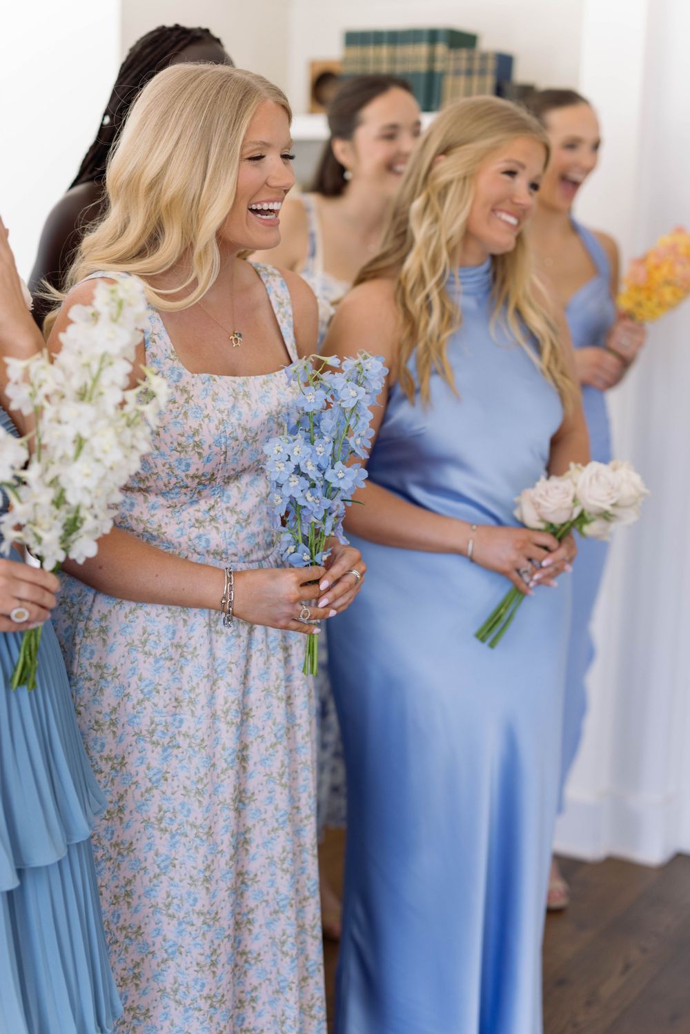 Bridesmaids wearing light blue dresses hold floral bouquets while laughing joyfully at an indoor wedding celebration.