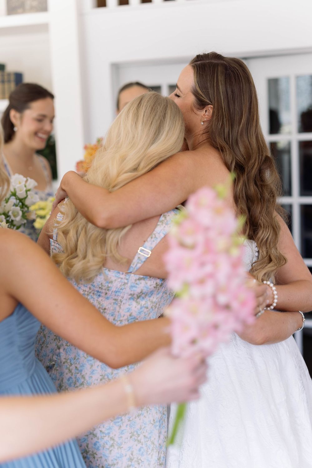 Emotional wedding moment as bridesmaids in pastel dresses embrace the bride while holding pink flowers.
