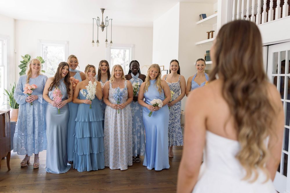 Bridesmaids in light blue dresses stand together indoors while looking at someone in a white wedding gown.
