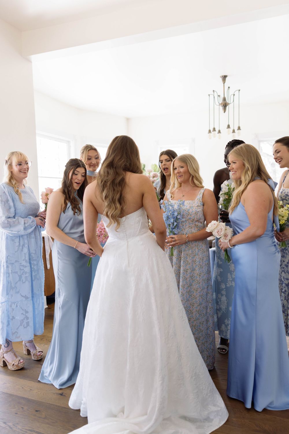 A wedding party in light blue bridesmaid dresses and a white wedding gown gather and chat in a bright indoor venue.