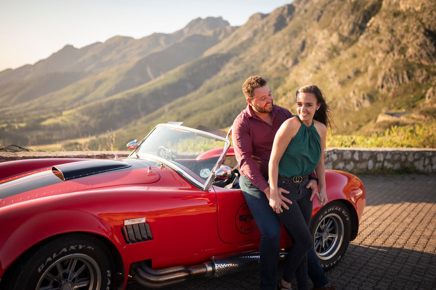 A couple shares a romantic moment while leaning against a vintage red sports car on a scenic mountain road at sunset.