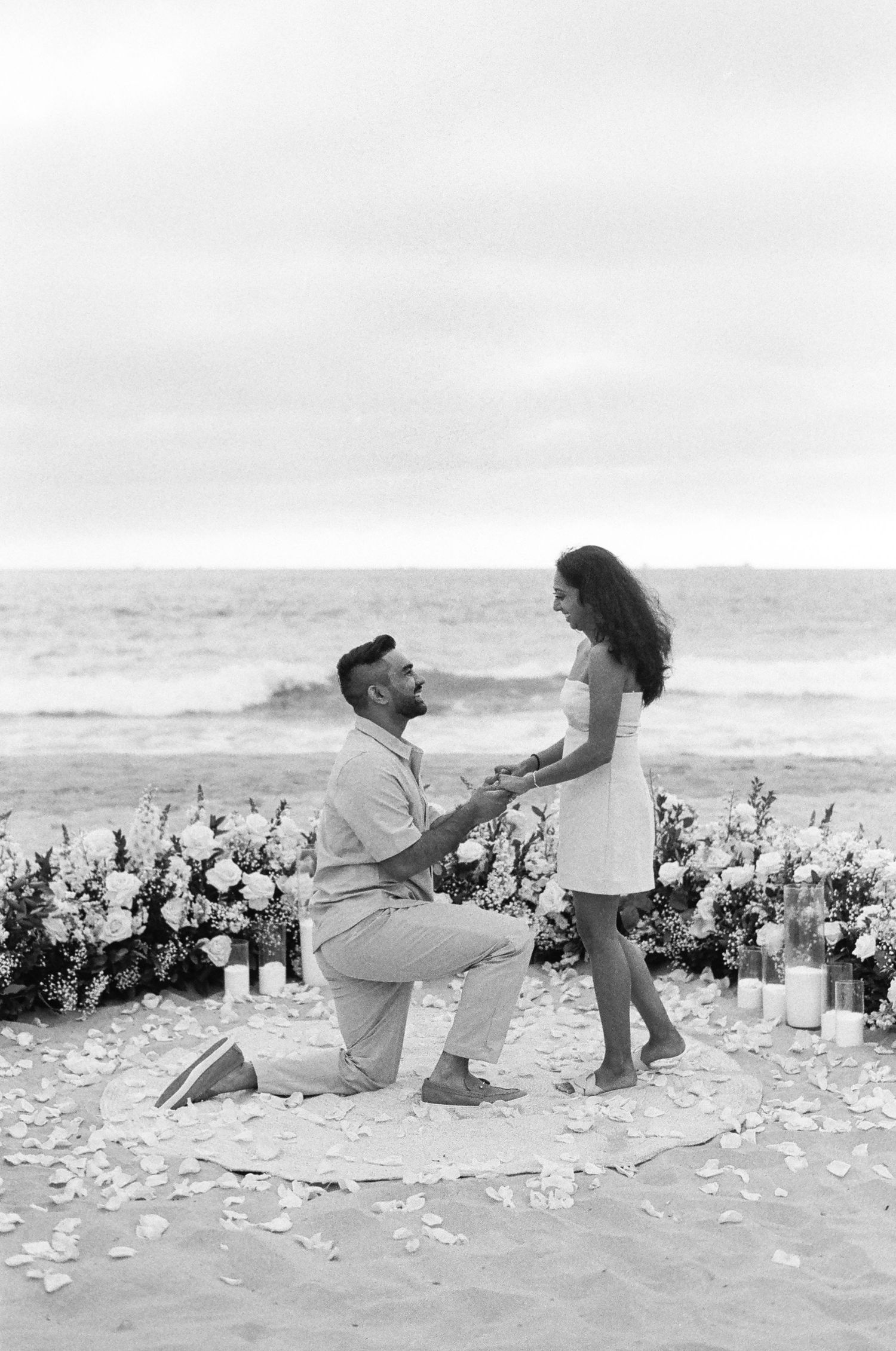 A romantic beach proposal setup with white floral arrangements and candles on the sand as a couple share their special moment.