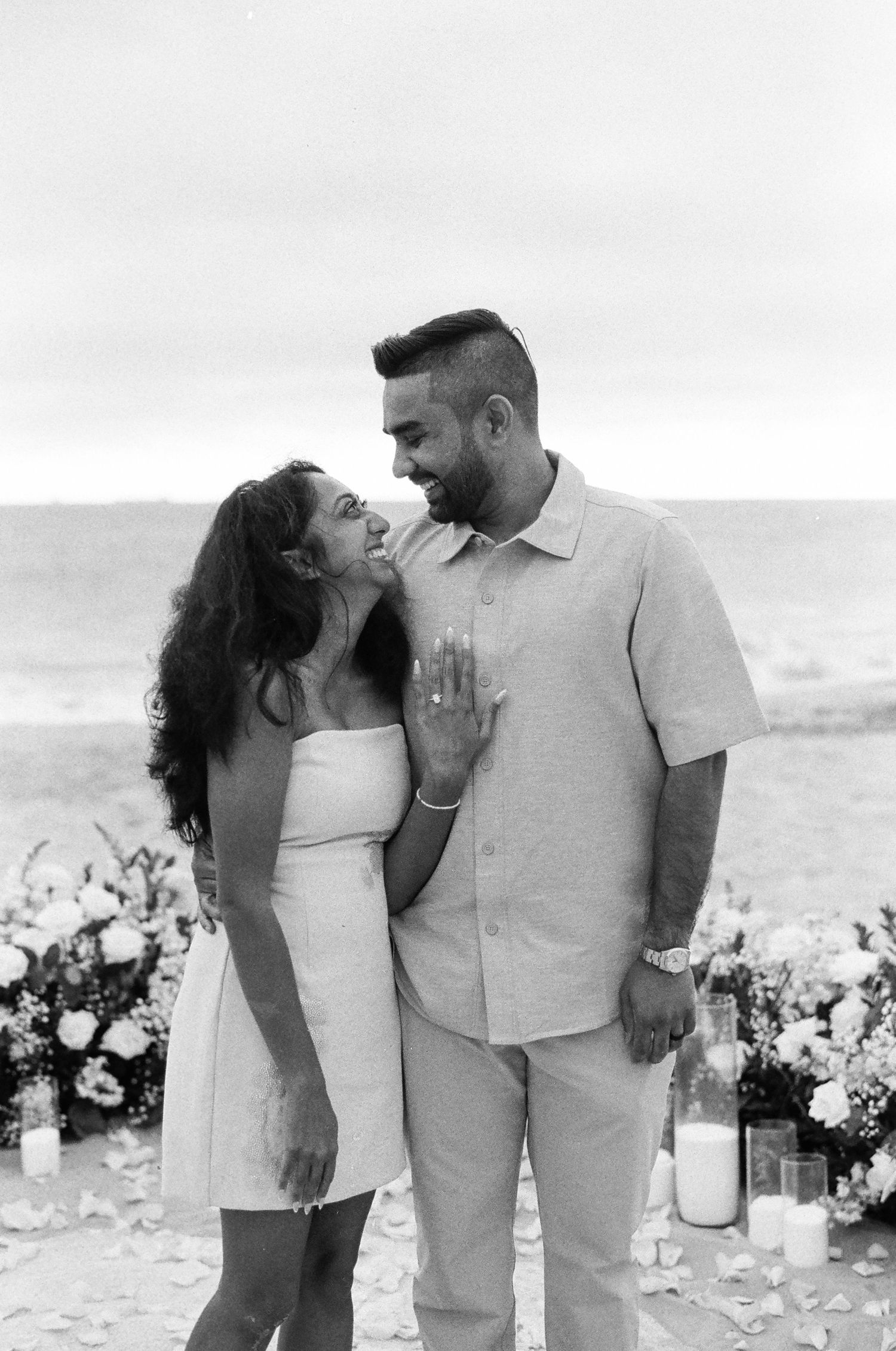 A romantic couple shares an intimate moment on the beach with floral decorations and candles at sunset.