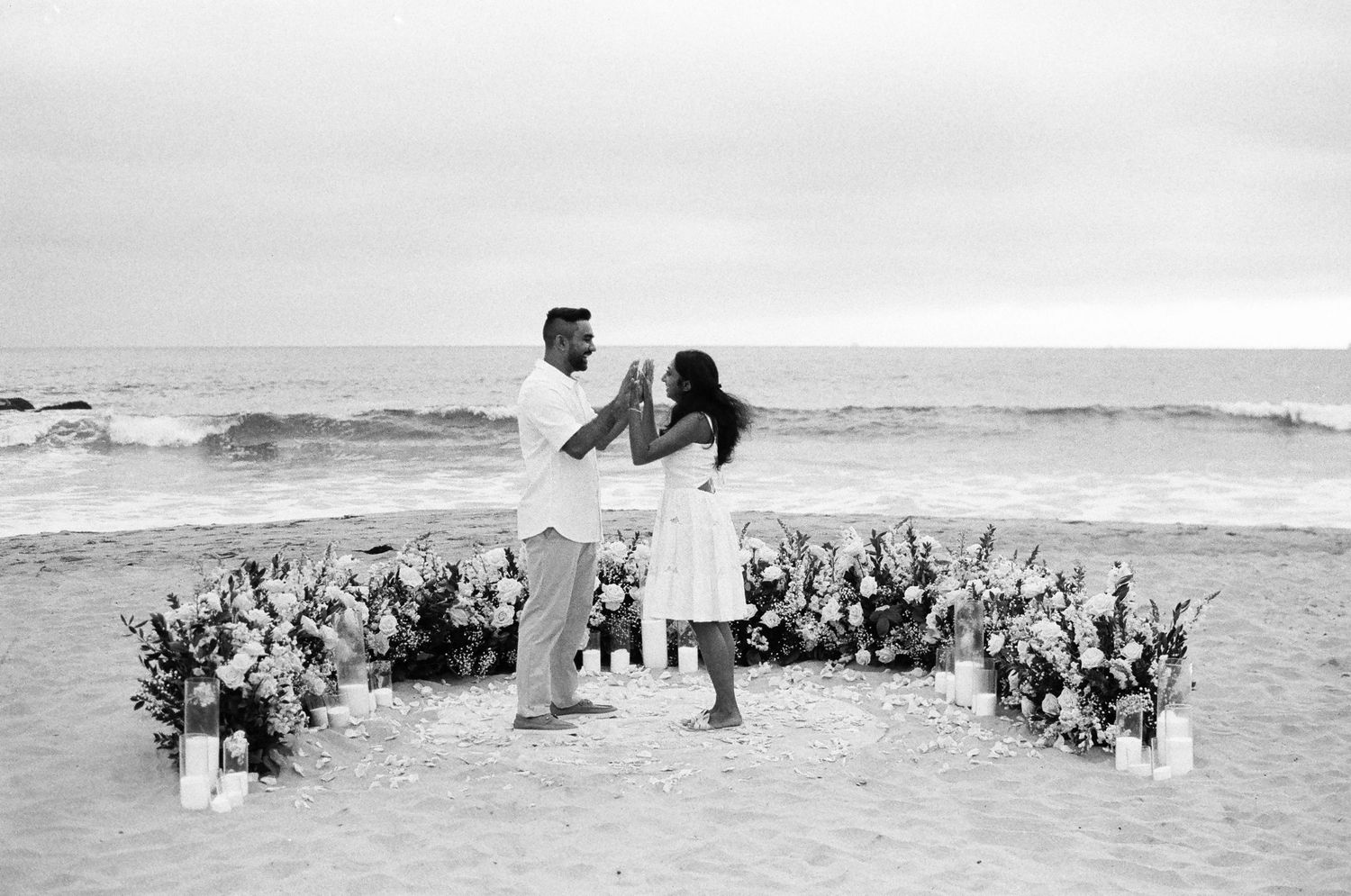 A couple embraces on a beach during an intimate ceremony surrounded by floral arrangements in black and white.