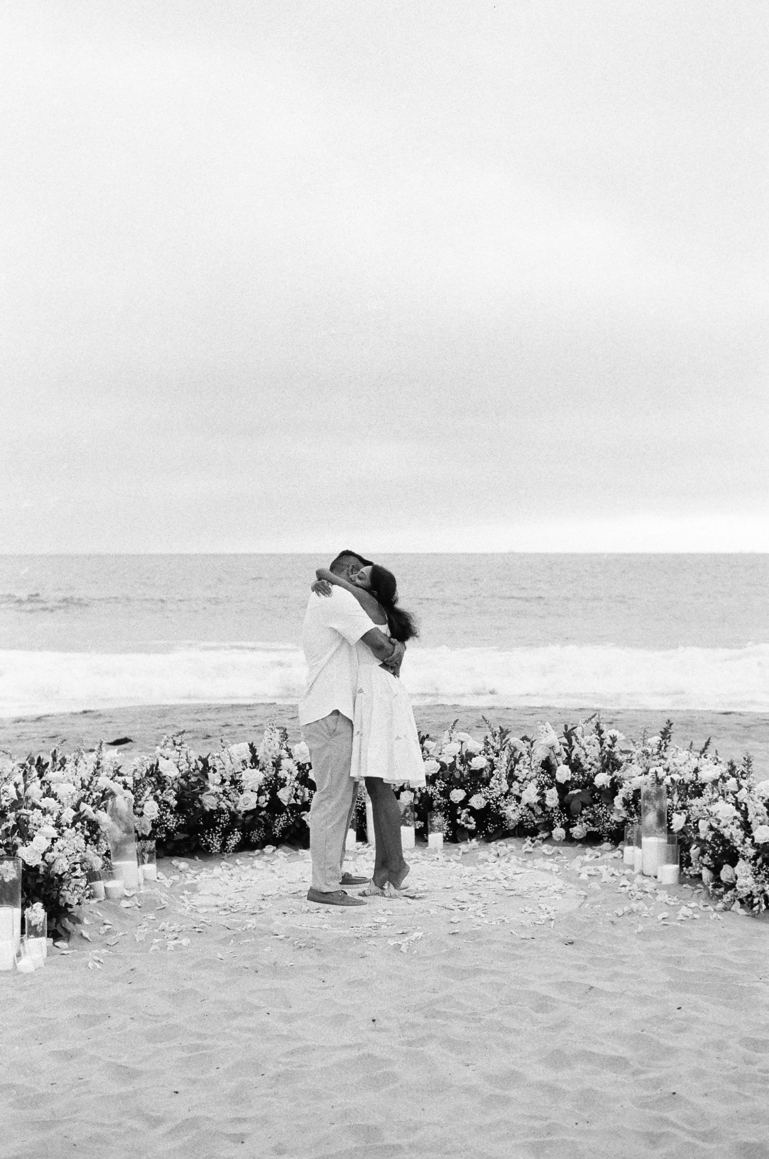 A romantic moment between two people surrounded by flowers on a beach with ocean backdrop.