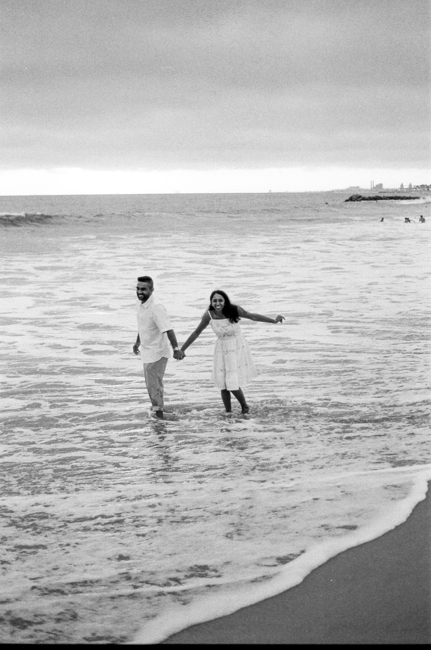 A couple holds hands while playing in the ocean waves along the shoreline in black and white.