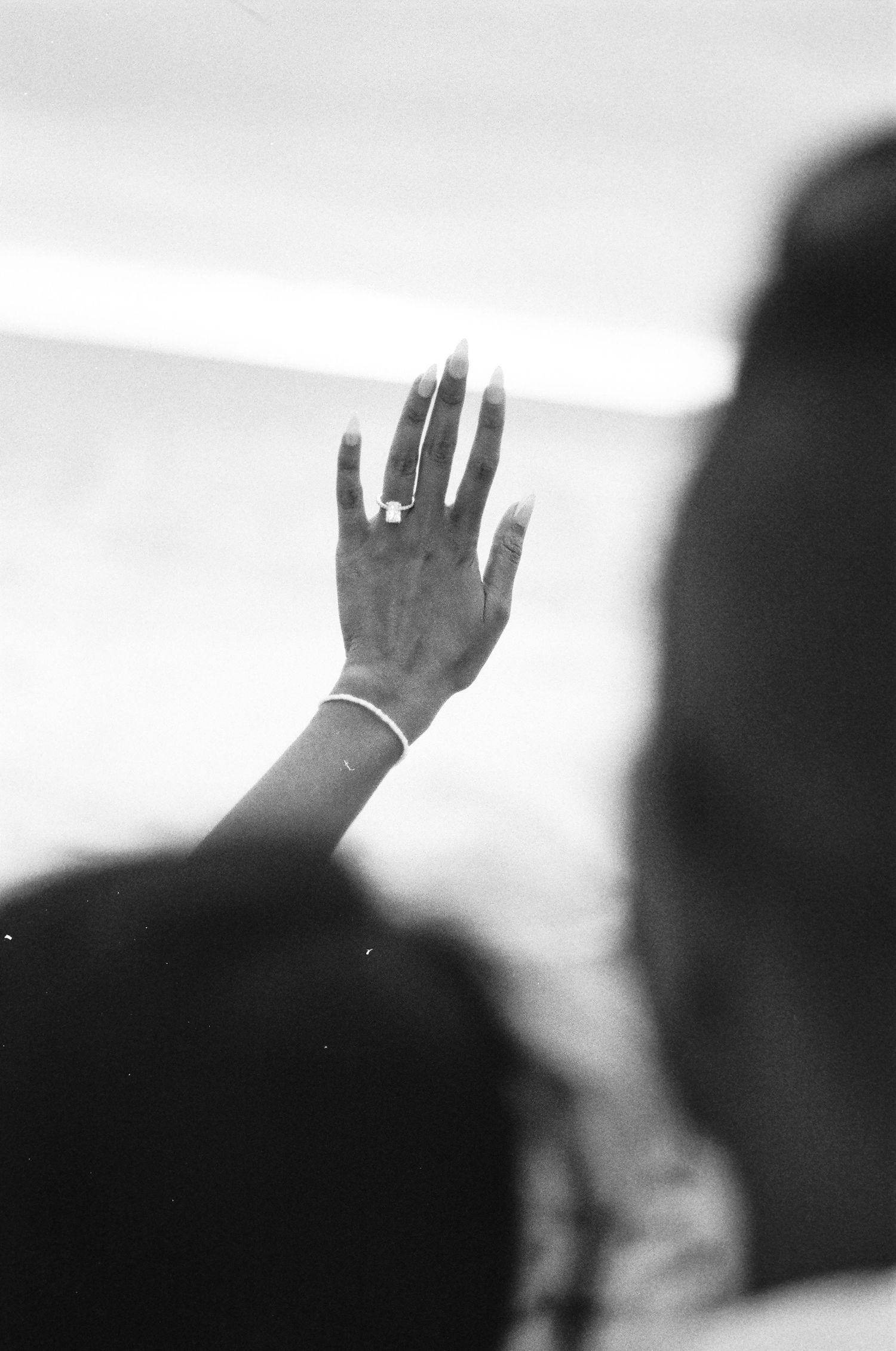 A close-up black and white photograph of a raised hand with a ring, silhouetted against a bright background.