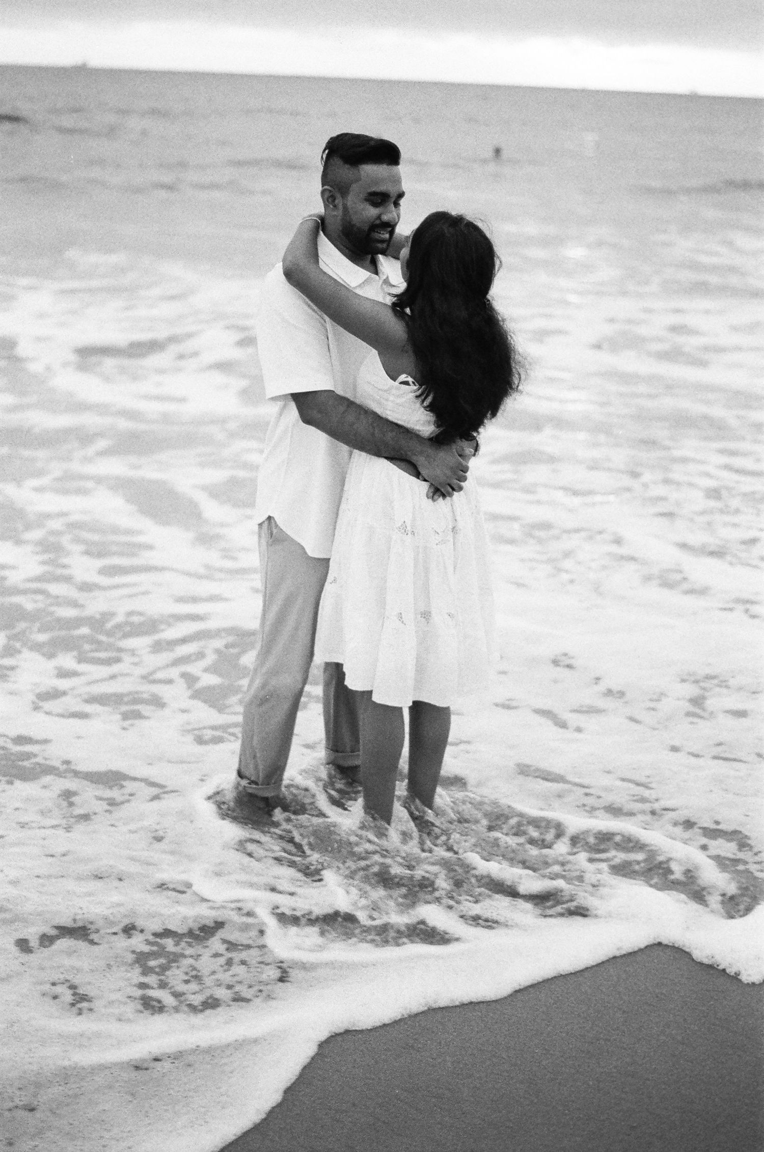 A loving couple embraces in the shallow ocean waves as water swirls around their feet.