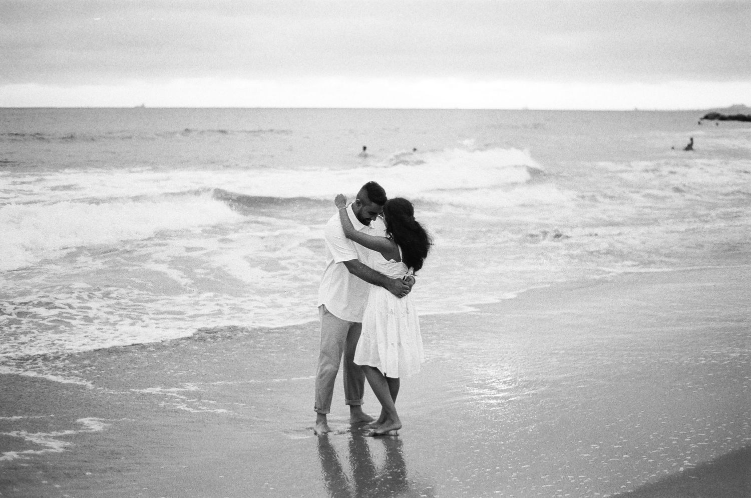 A romantic black and white photograph of two silhouettes embracing on a misty beach with waves crashing behind them.