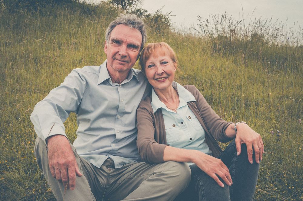 An elderly couple sits together on a grassy hillside during a sunny afternoon, captured in a warm vintage-style photo.